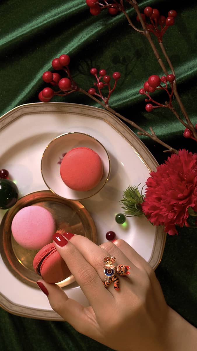 A hand with a tiger ring with a butterfly and floral elements, holds a pink macaron, surrounded by flowers and berries.