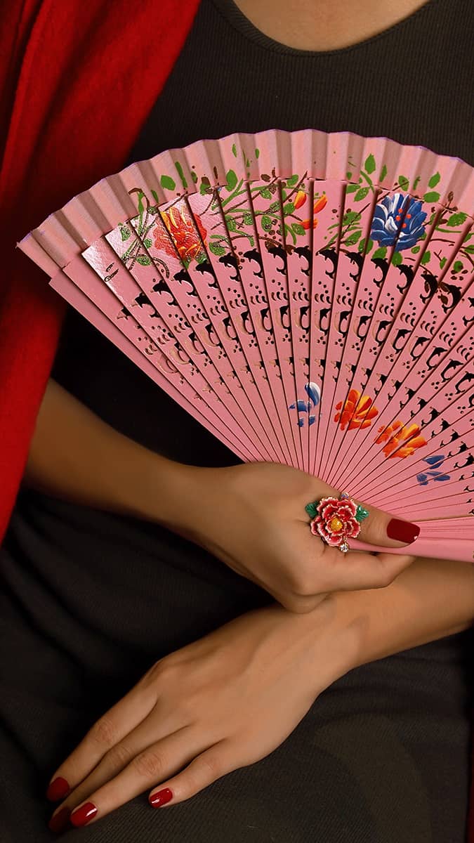 A beautifully decorated pink fan held by a hand, adorned with a beautiful floral ring and polished red nails.