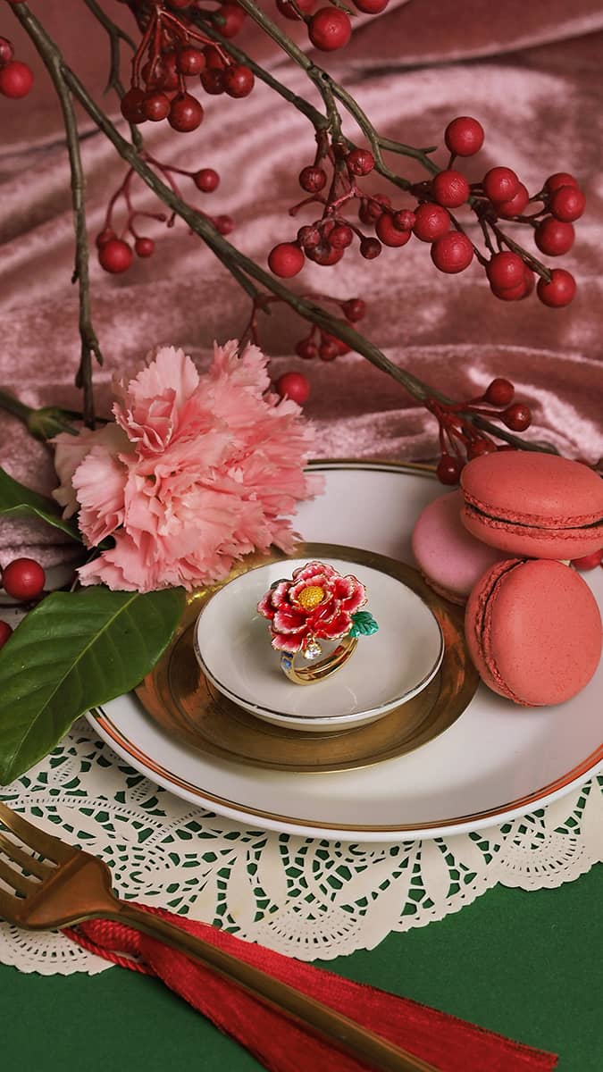 A delicate pink carnation, a golden ring with a floral design, pink macarons, and decorative red berries on a lace doily.