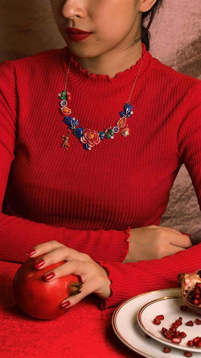 A woman in red sweater wearing a stunning statement floral necklace with a small tiger and butterfly charm while holding a pomegranate.

