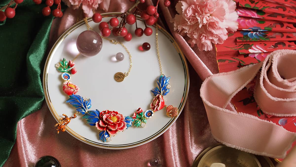 A vibrant statement necklace featuring intricately crafted floral designs in red, blue, with small tiger and butterfly charm, is displayed on a white circular plate surrounded by decorative items.