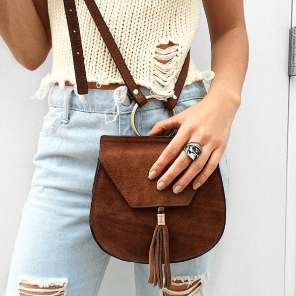 A woman in causal outfit wearing a stylish skull ring on her hand while resting on a brown bag.