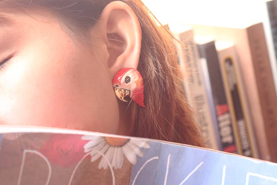 Unique colorful bird earrings worn by a woman, surrounded by books.