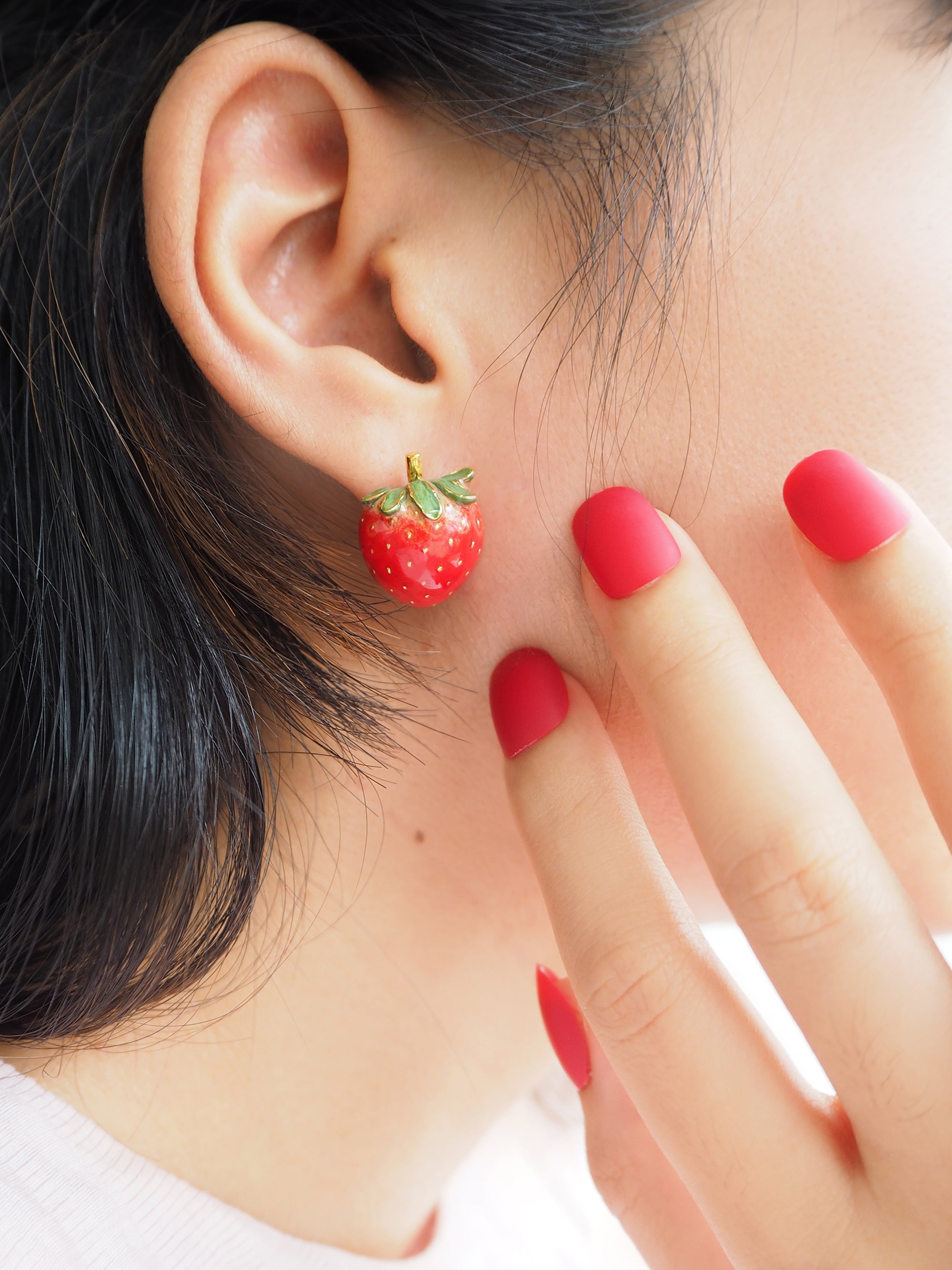 Red strawberry-shaped earring worn by a person touching their face with a red-manicured hand.