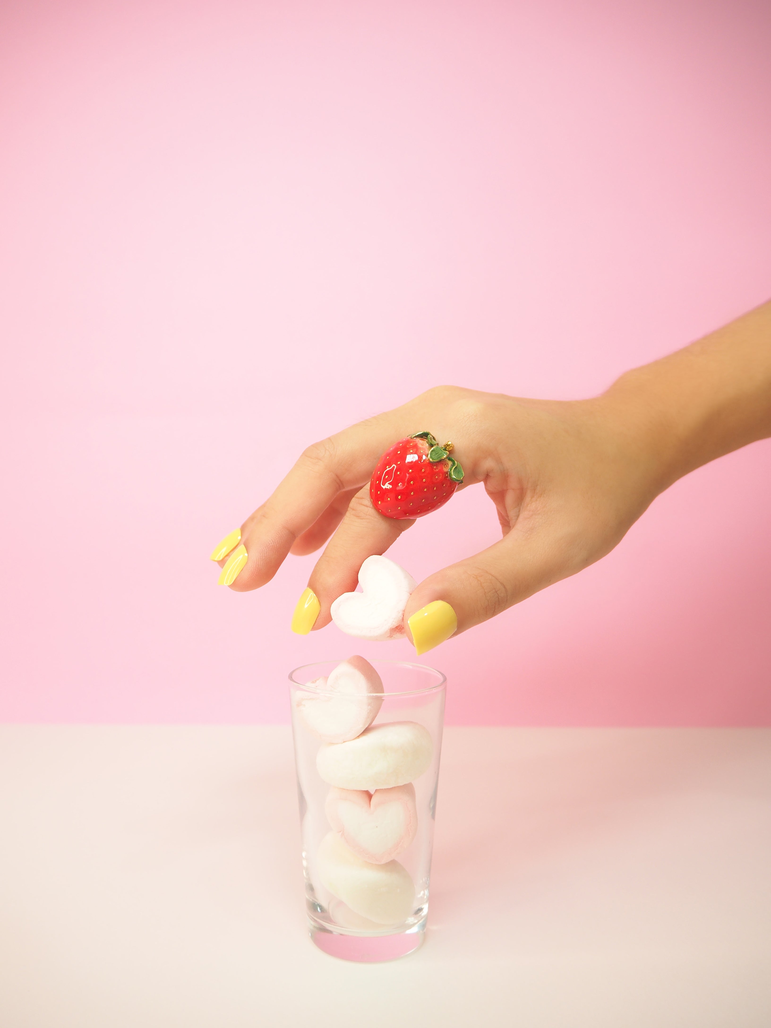 A hand with a strawberry ring drops heart-shaped marshmallow into a glass.