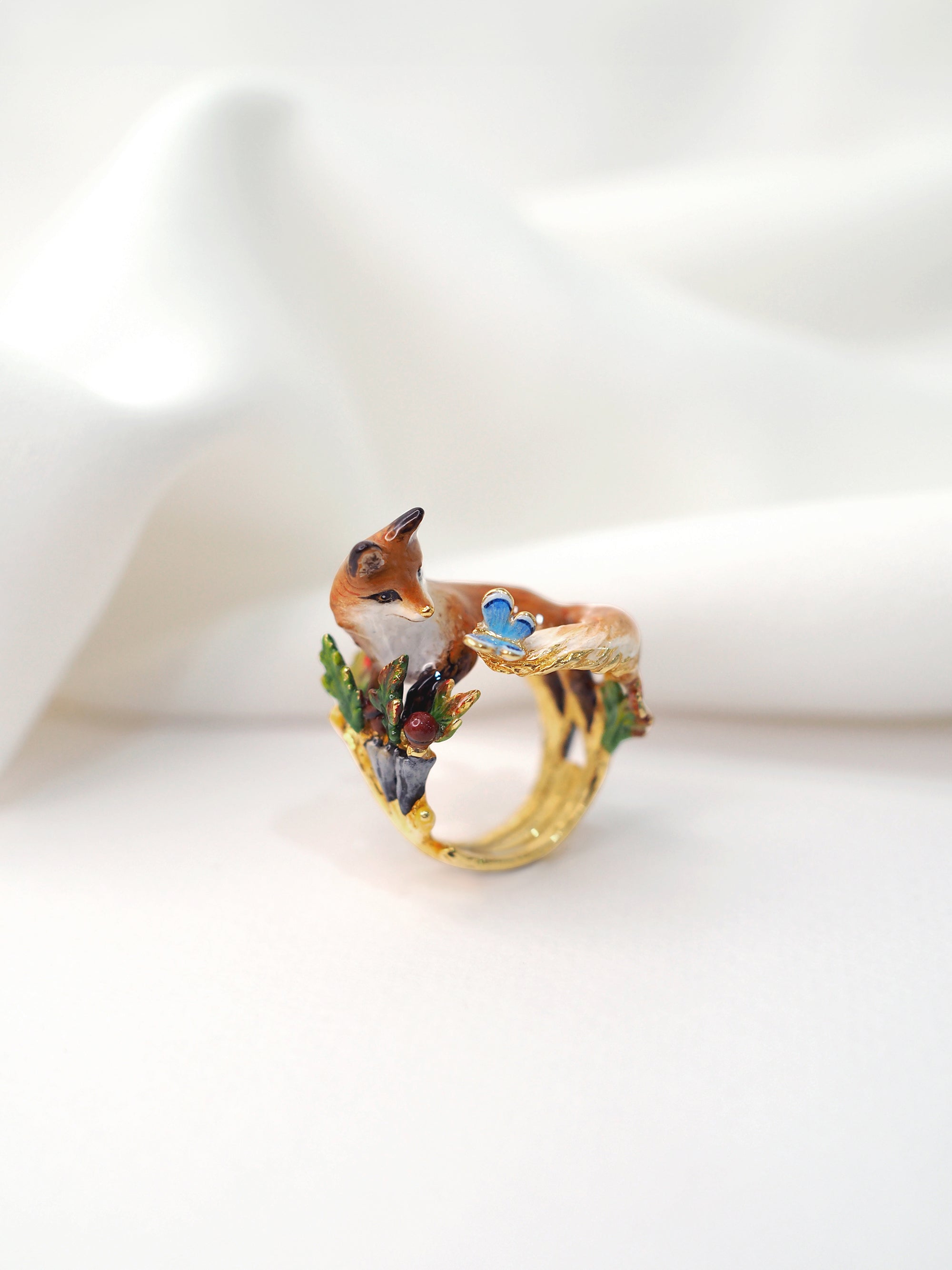 A beautifully crafted gold ring featuring a fox and a butterfly, adorned with colorful enamel details placed on white fabric.