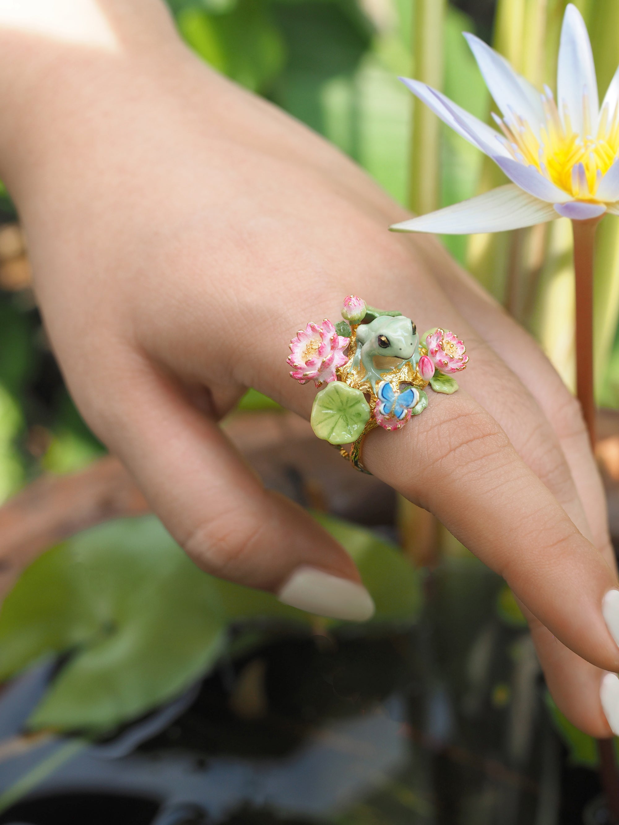 A nature-inspired ring showcasing a frog and pink flowers, worn on a finger.