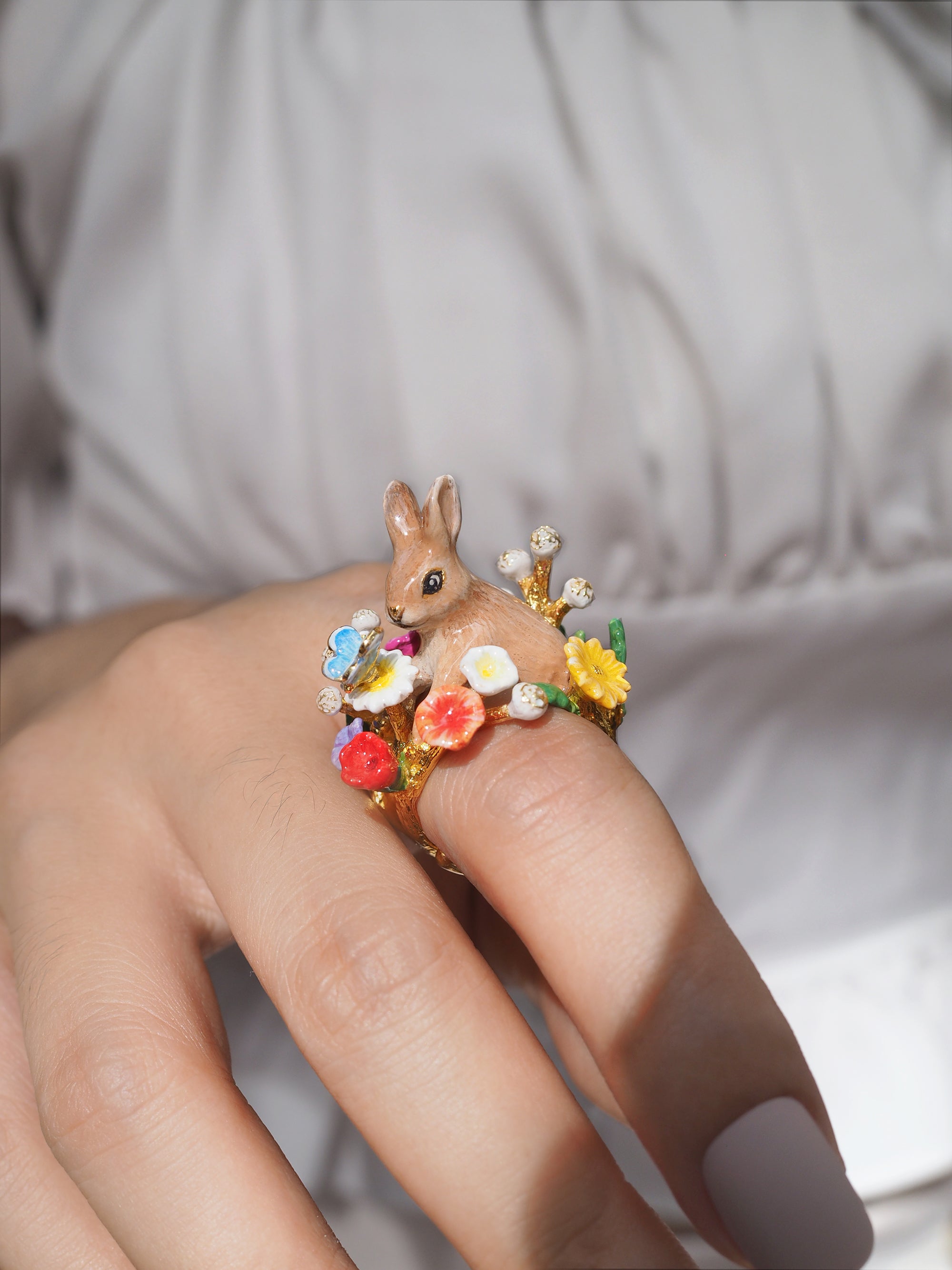 Close-up of a hand showcasing a ring featuring a rabbit surrounded by colorful flowers and a blue butterfly