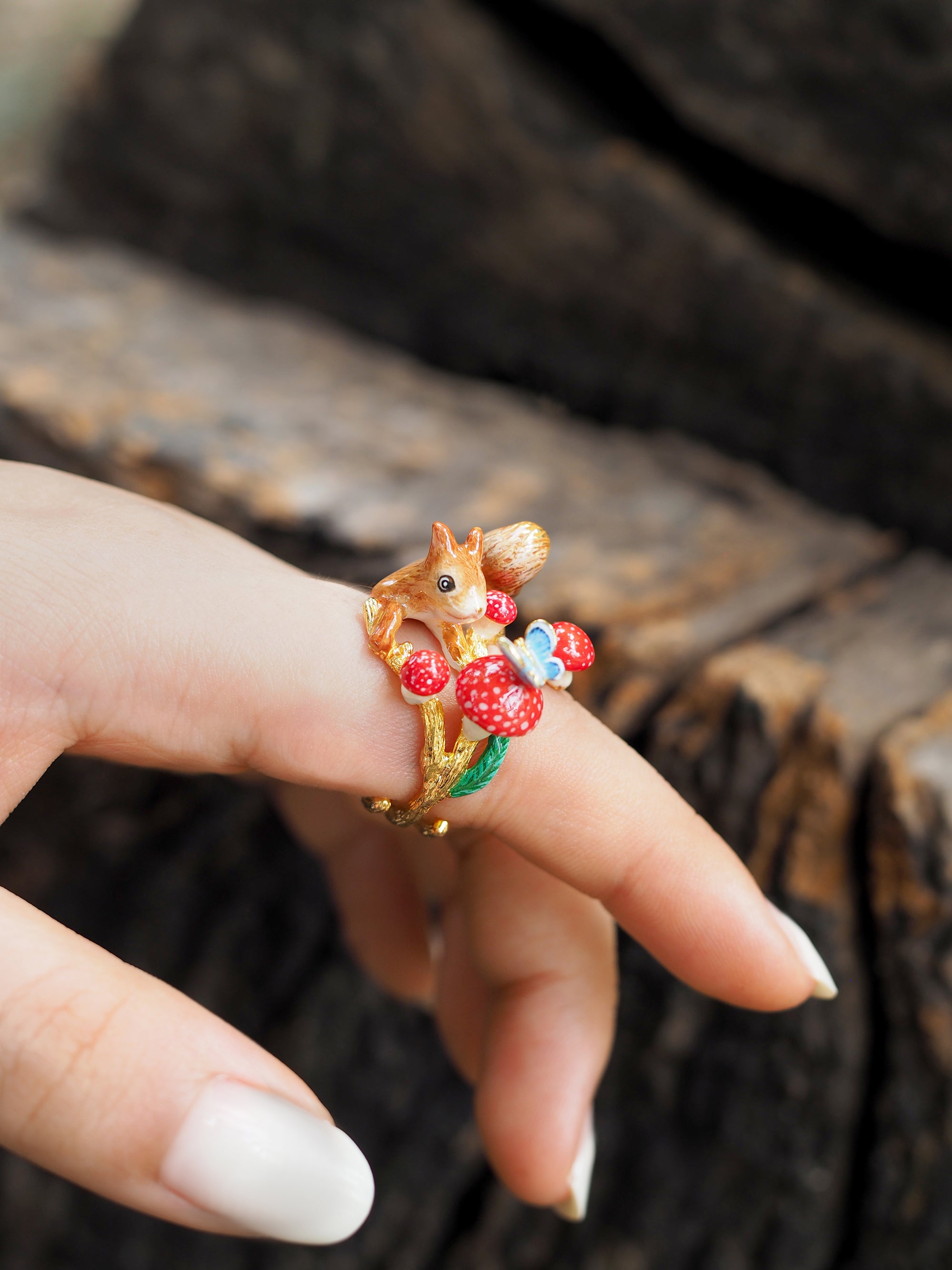 Close-up of a hand adorned with a gold ring featuring a squirrel, mushrooms, and a blue butterfly design in natural setting.