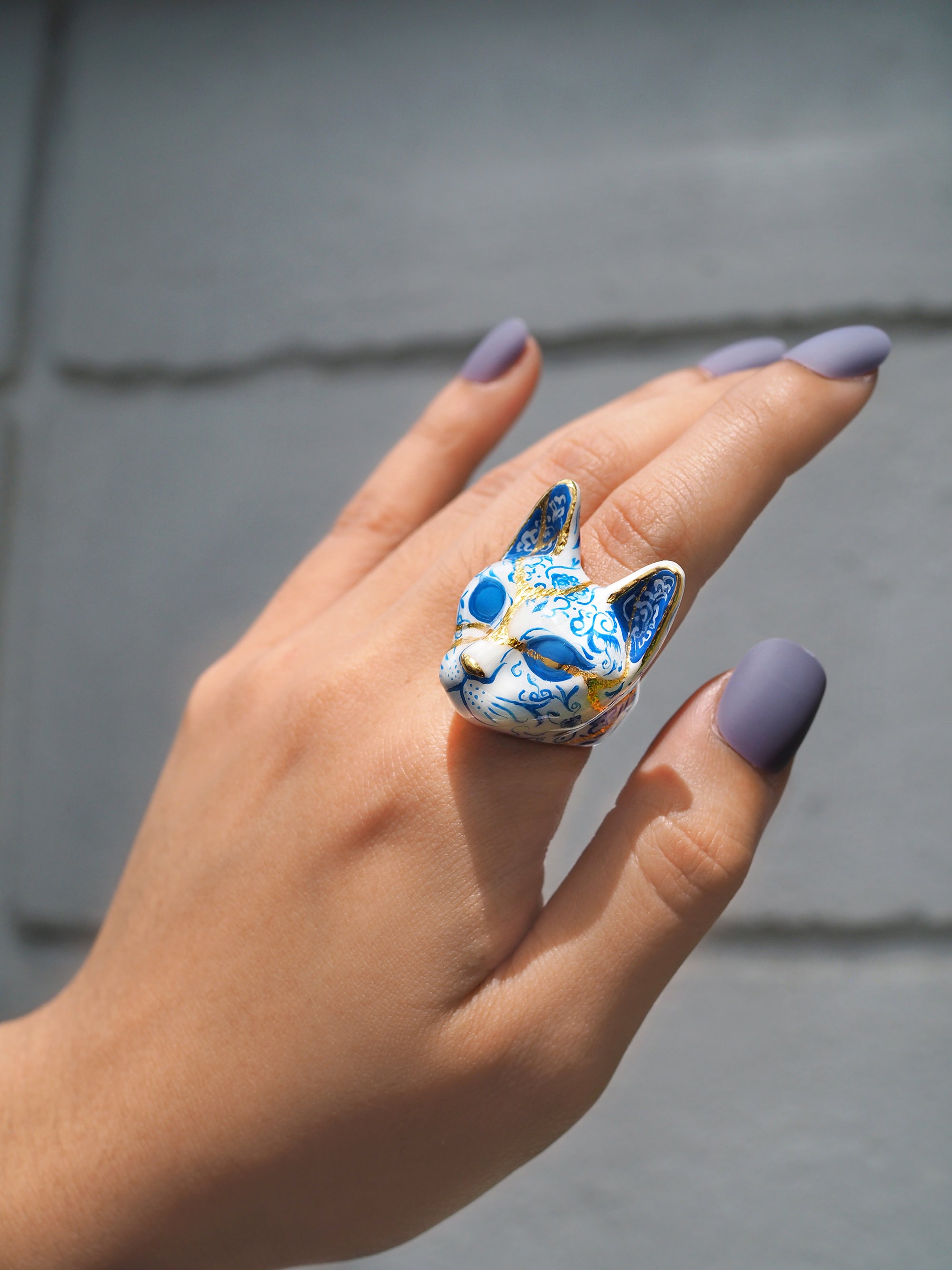 A hand with lavender nails wearing a blue and white cat ring, detailed with porcelain patterns.