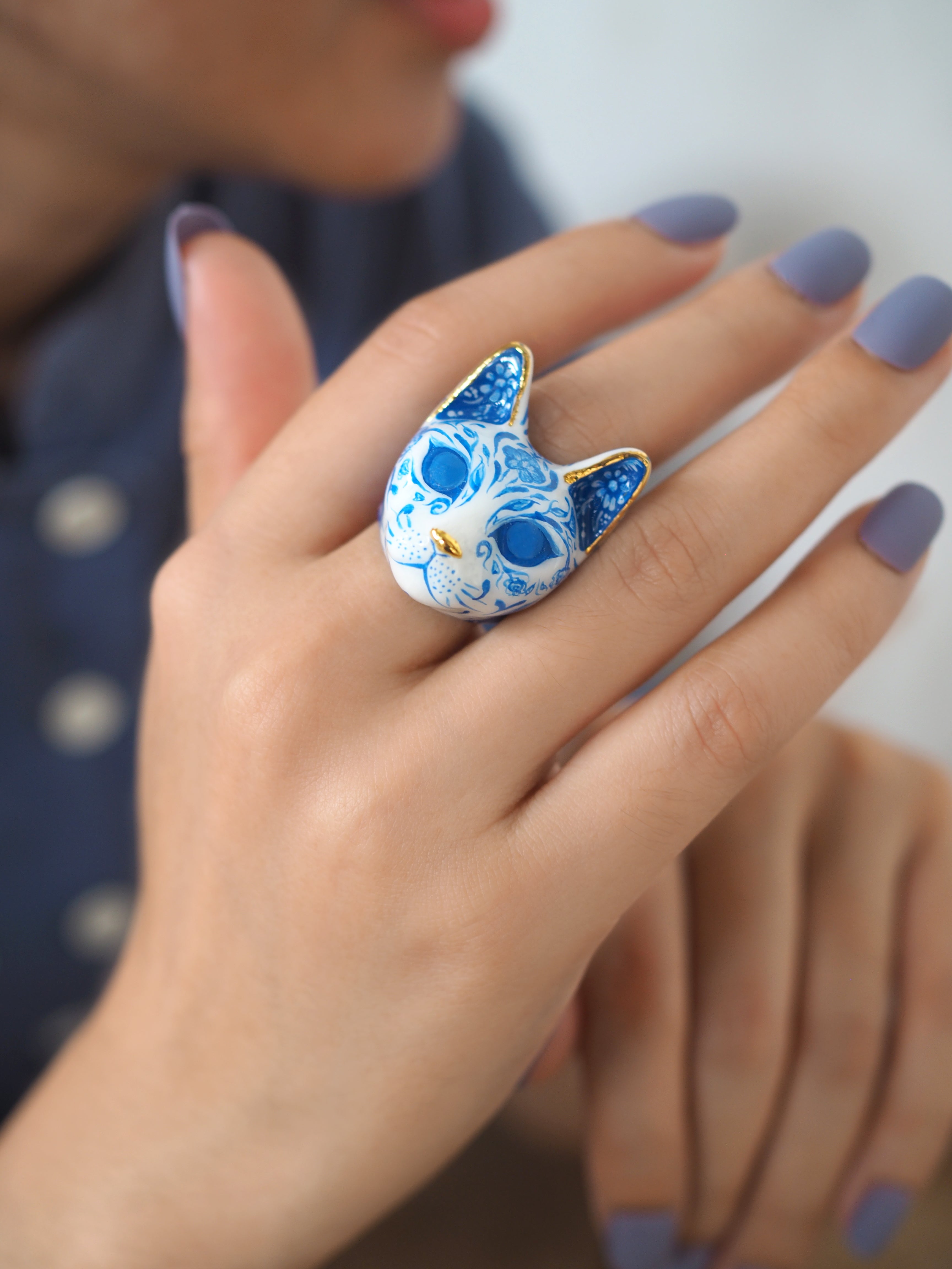 Close up of a hand with lavender nails showcases a blue and white cat ring, detailed with porcelain patterns.