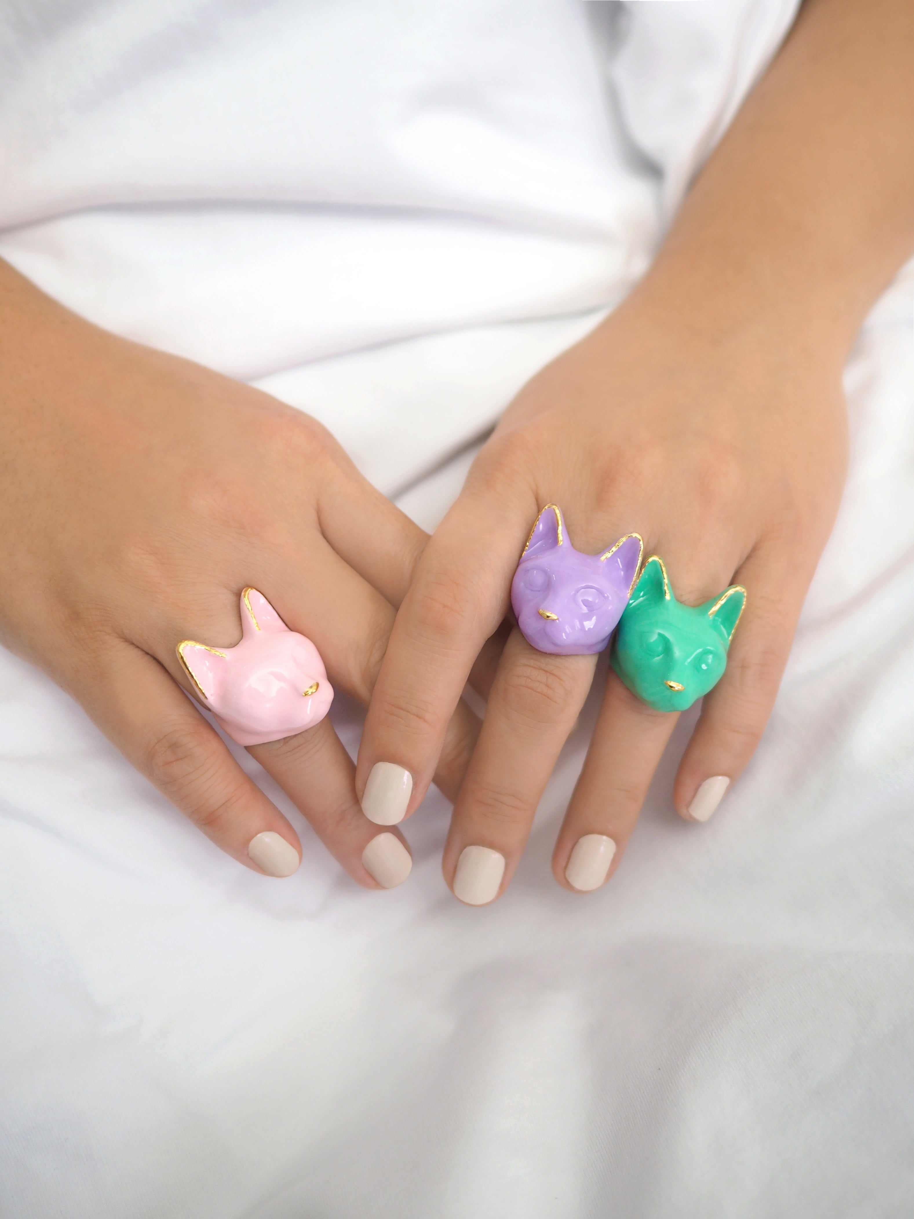 Three colorful cat-shaped rings—pink, lavender, and green—are worn on a person's manicured fingers against a white background.