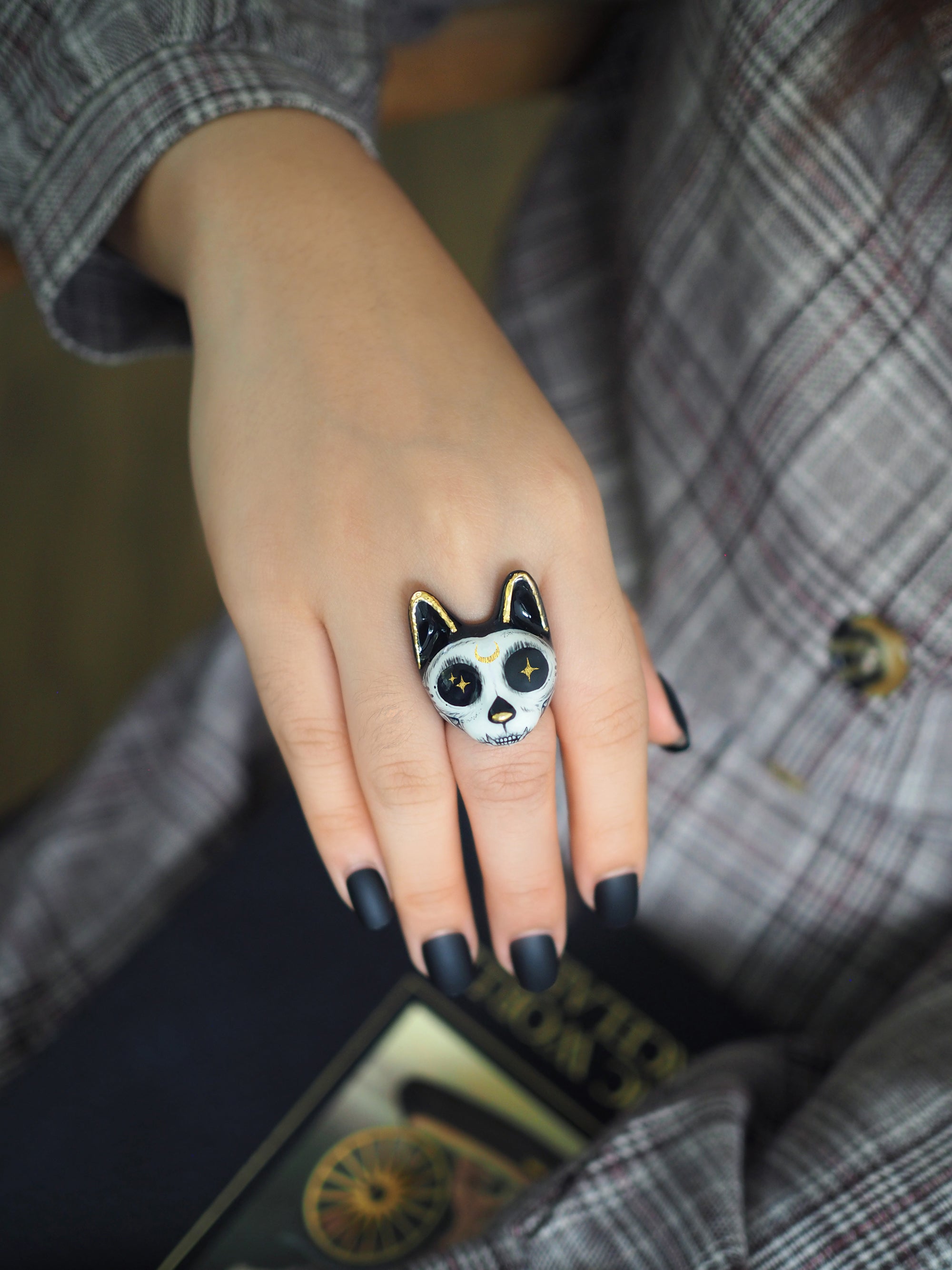 A close-up of a hand wearing a unique black and white cat ring, featuring prominent ears and large, starry eyes rests against a patterned blazer.