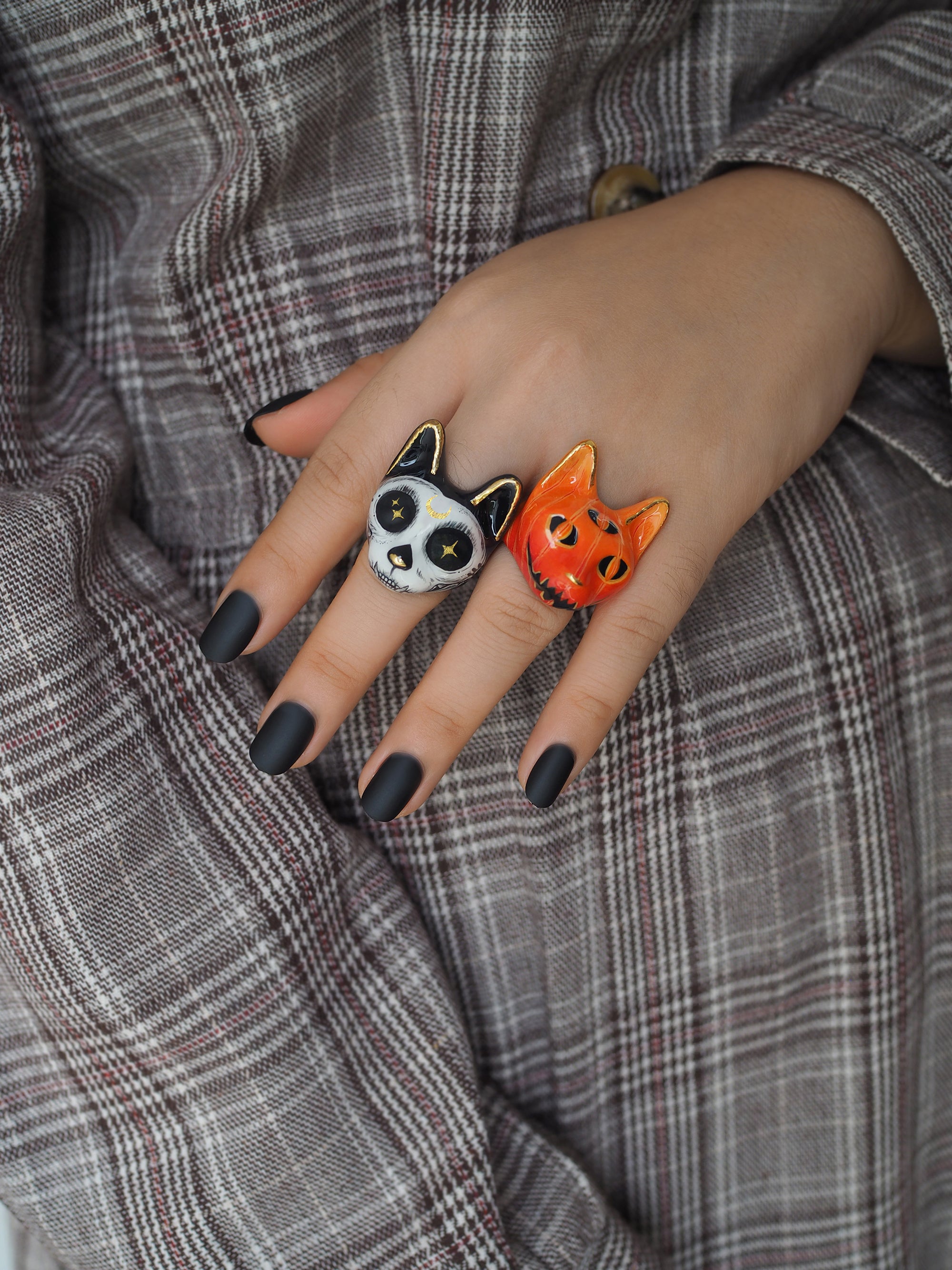 Two Halloween-themed cat rings on a hand, the left ring has a black and white cat face with skull design, while the right features an orange cat with Jack-o'-lantern design, set against a plaid fabric background.