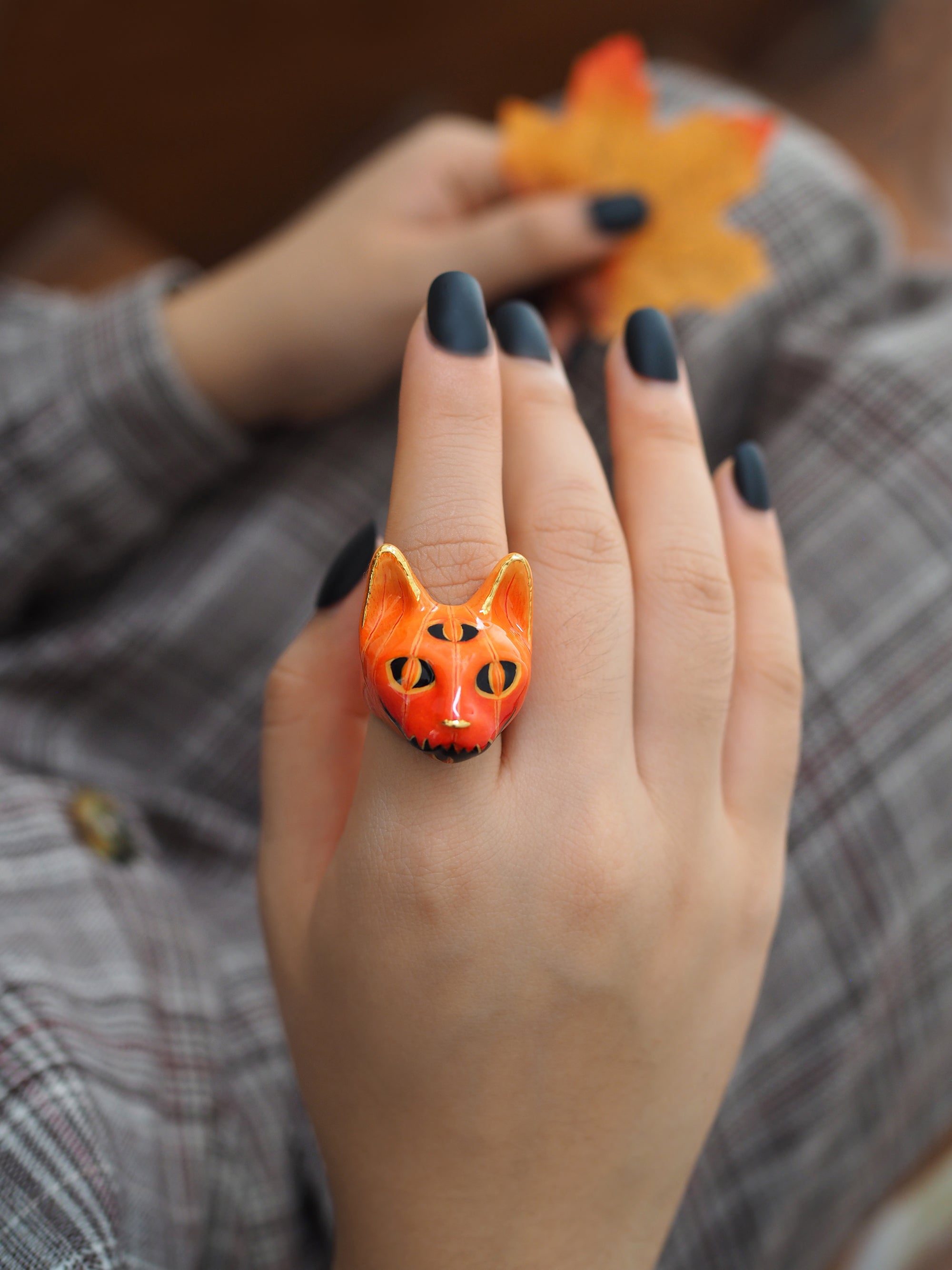 Halloween-themed ring featuring an orange cat's face, worn on a hand with dark-painted nails while another hand holding an autumn leaf.