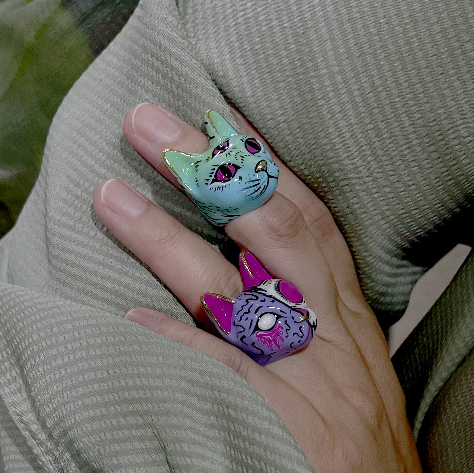 Close-up of a hand adorned with a vibrant green cat ring with a third eye design, and a purple cat ring with a zombie skull design.