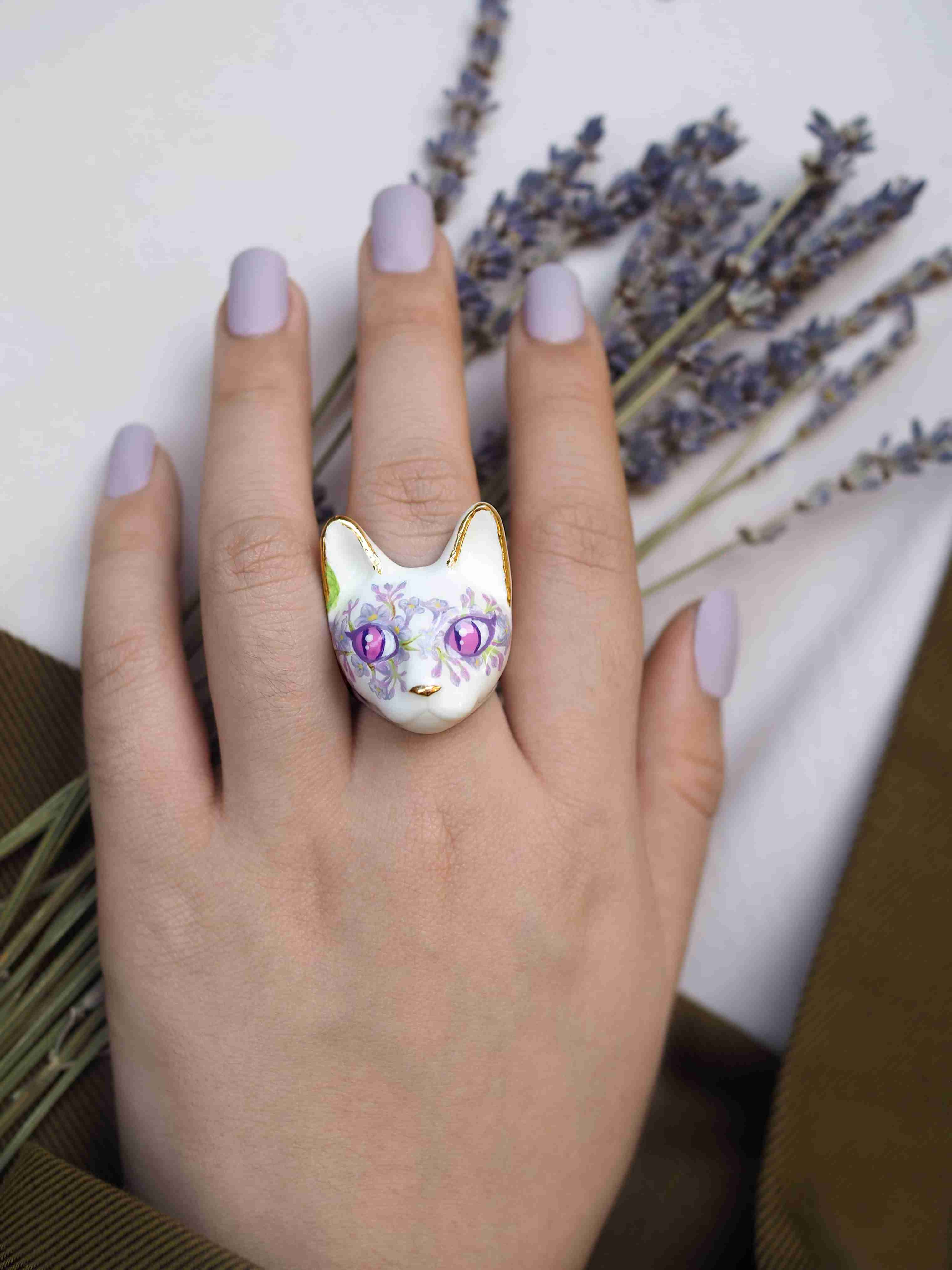 A hand with lilac nails wearing a unique cat ring with floral pattern, surrounded by dried lavender sprigs.