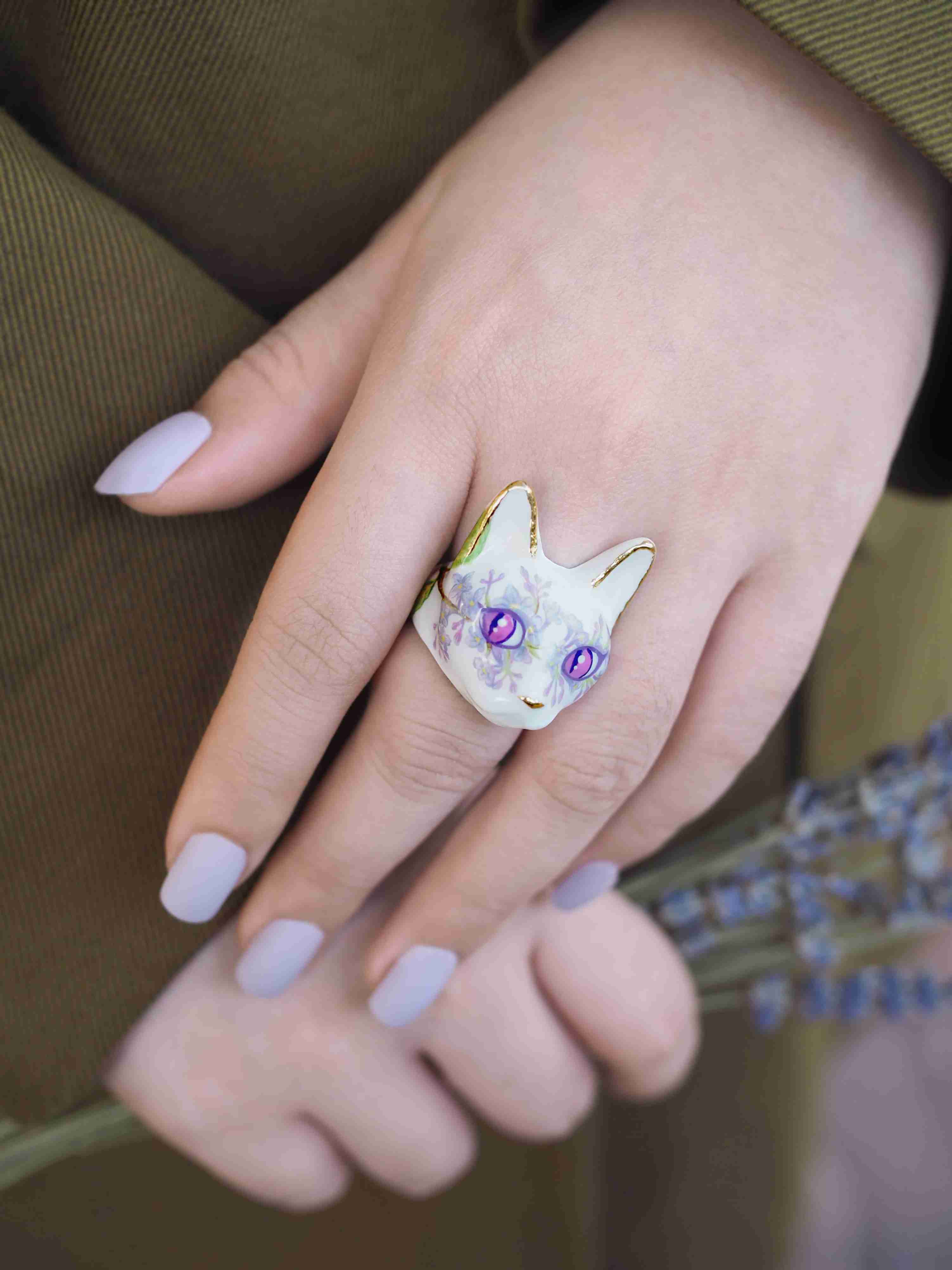 A hand with light purple nails wearing a whimsical cat face ring, featuring purple eyes and floral lilac pattern.