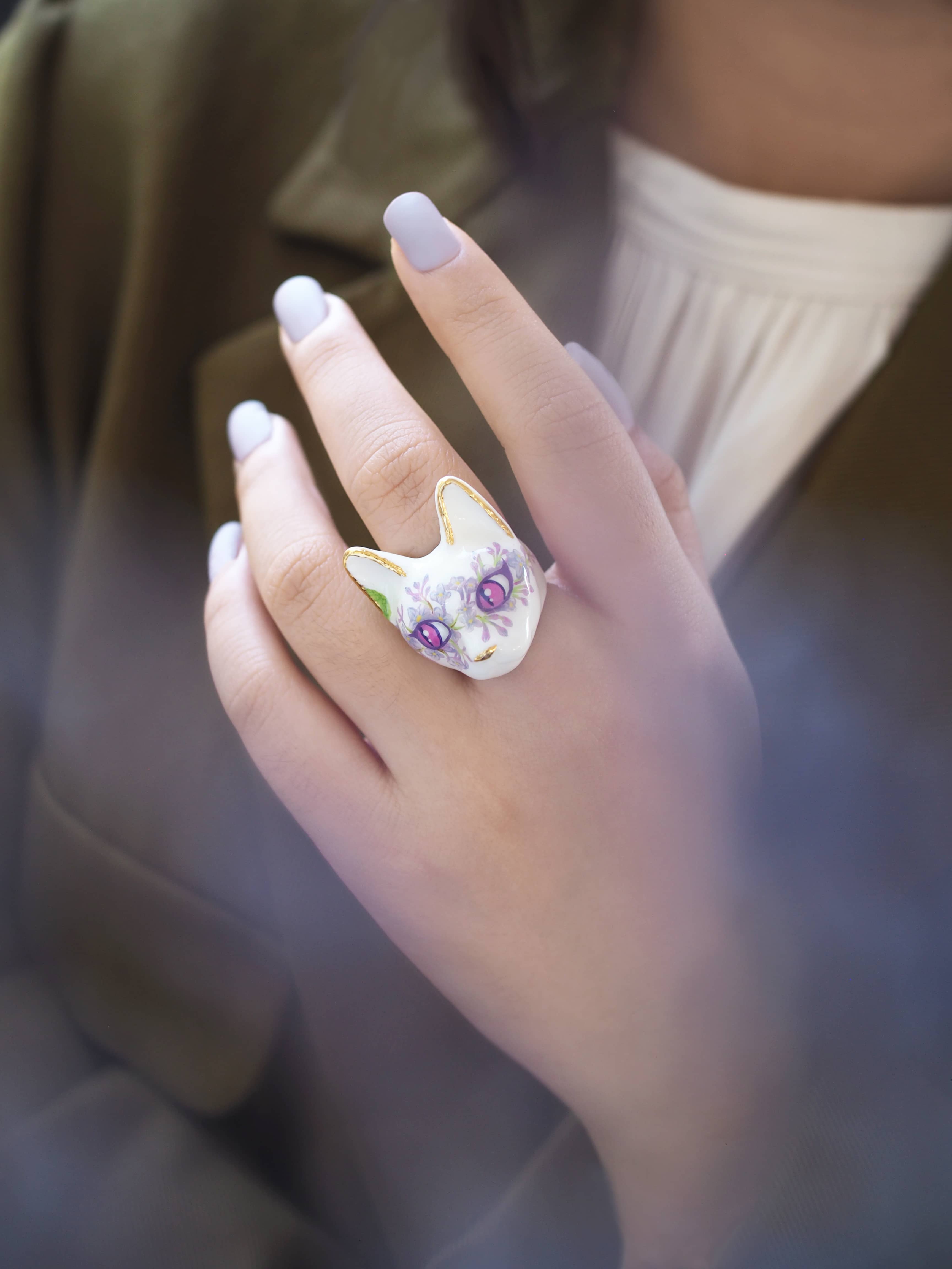 A hand with light purple nails wearing a whimsical cat face ring, featuring purple eyes and floral lilac design.