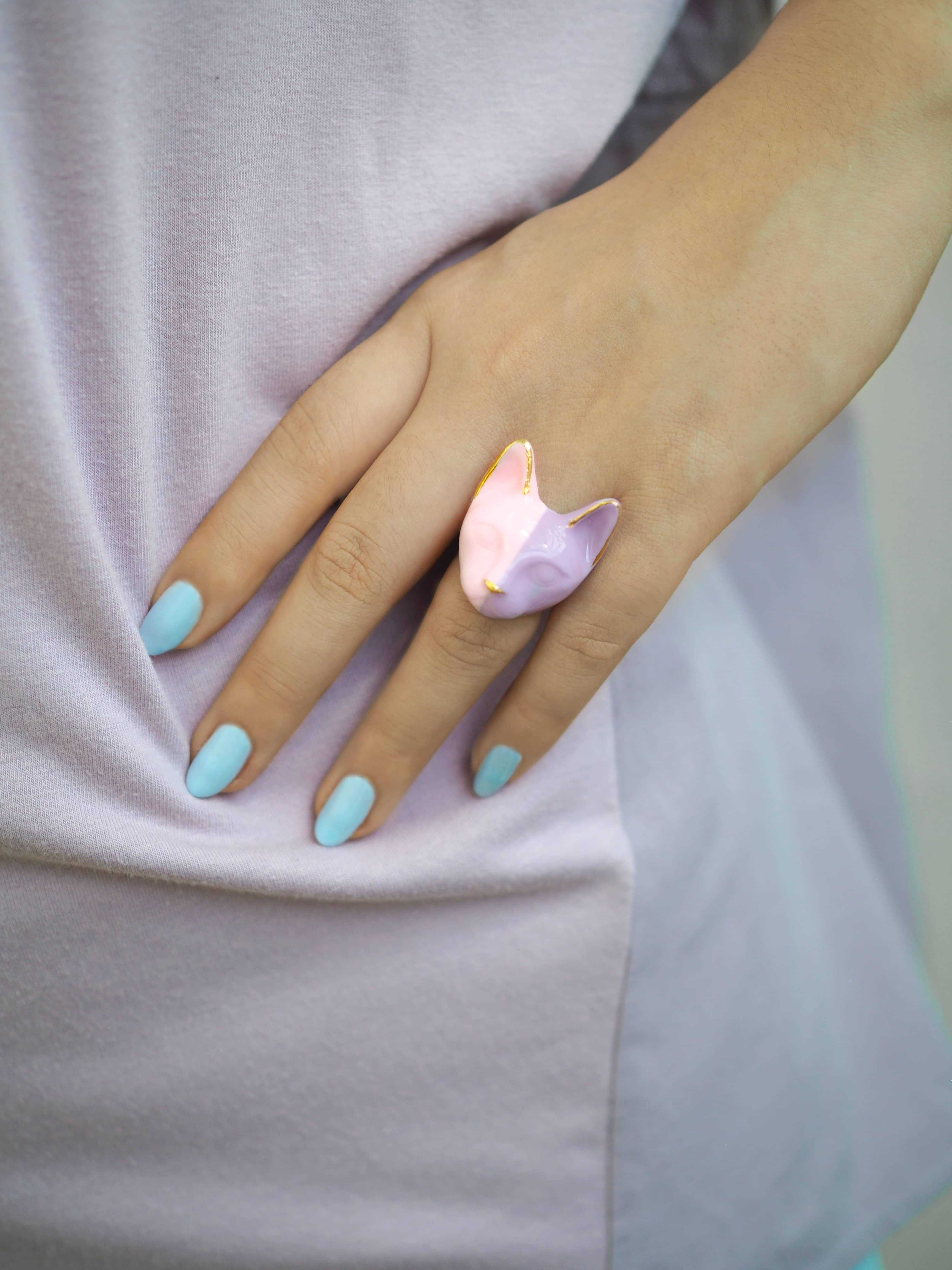 A hand showcasing a pastel purple and pink cat face ring with light blue nails.