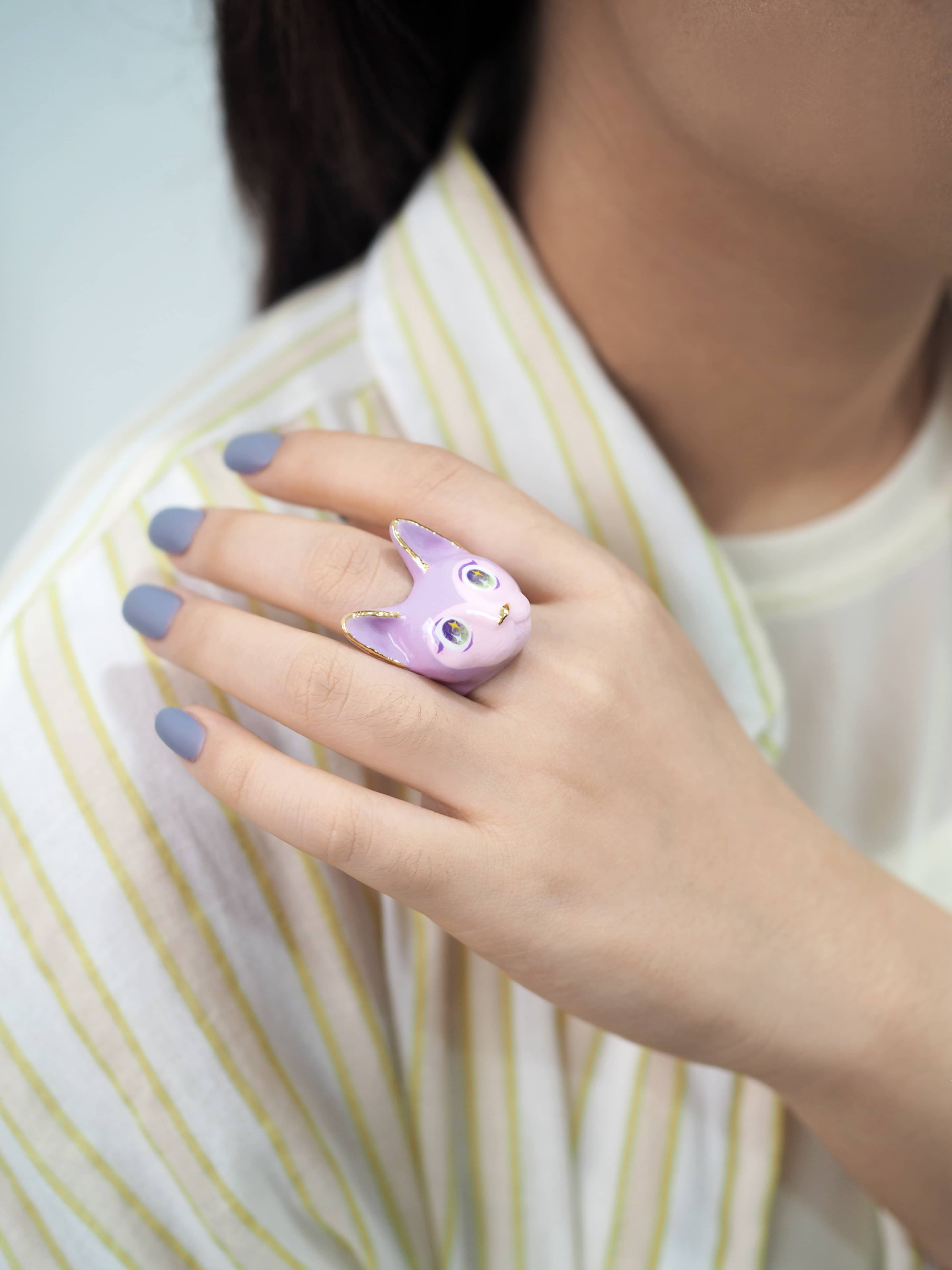 A hand with lavender nails wears a cute purple cat ring, resting gently against a striped shirt.