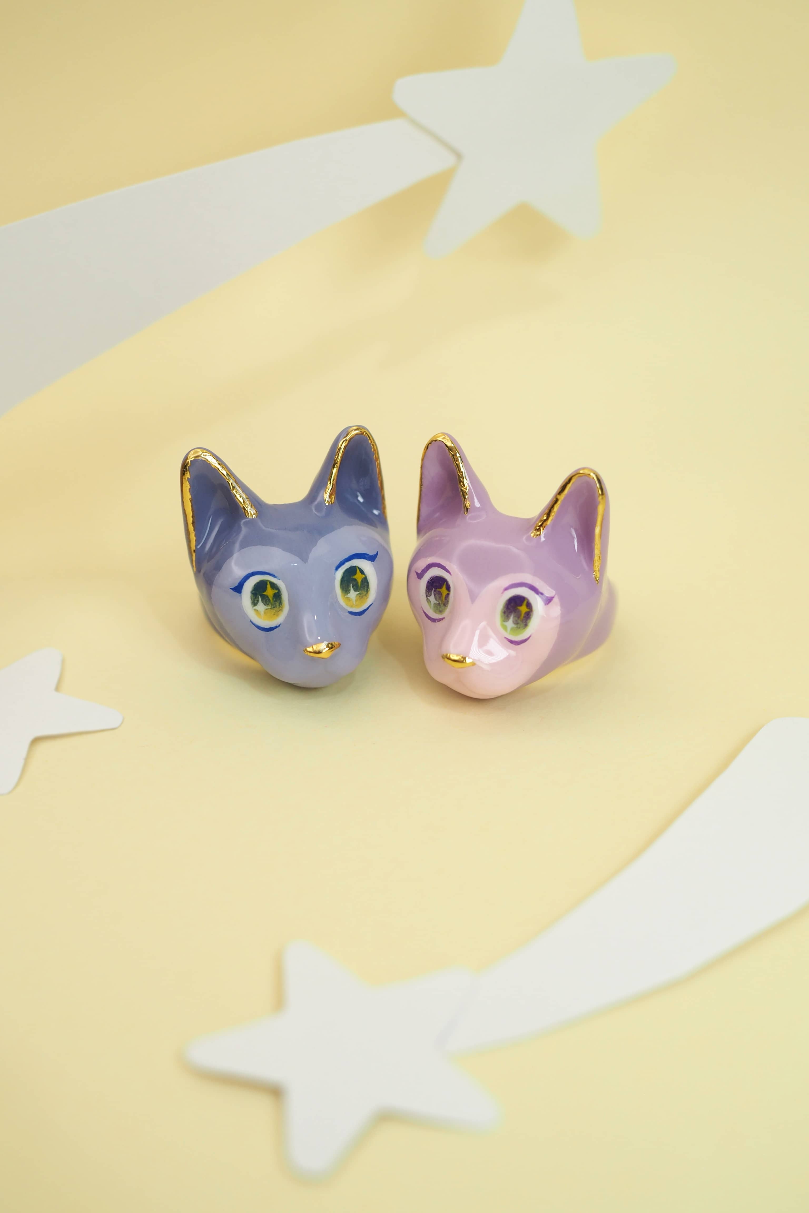 Two whimsical cat rings in purple and pink, each with big eyes placed on a soft yellow background with stars.