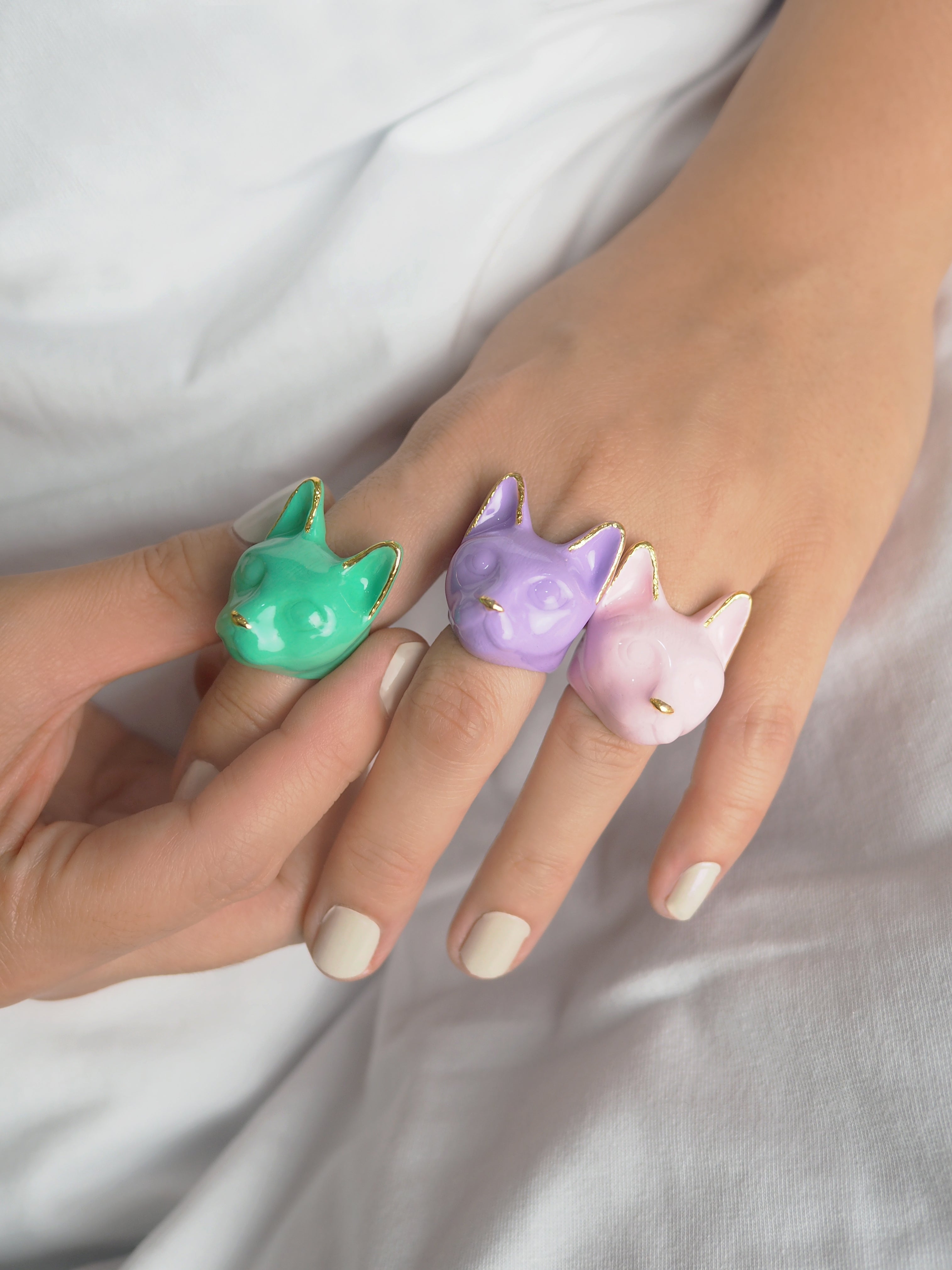 Three colorful cat-shaped rings—pink, lavender, and green—are worn on a person's manicured fingers against a white background.