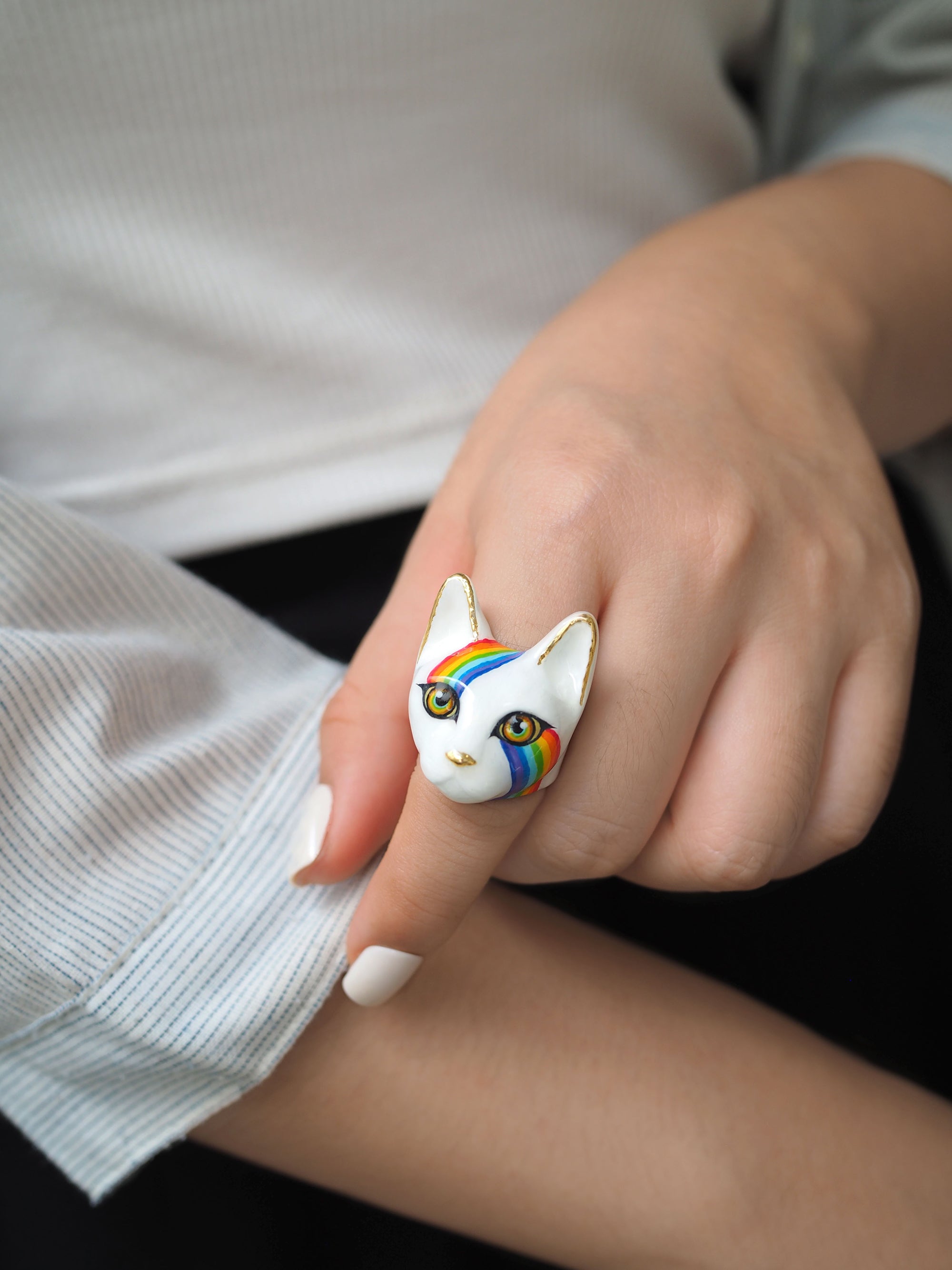 A close-up of a hand with a whimsical cat ring featuring a white base with rainbow stripes.