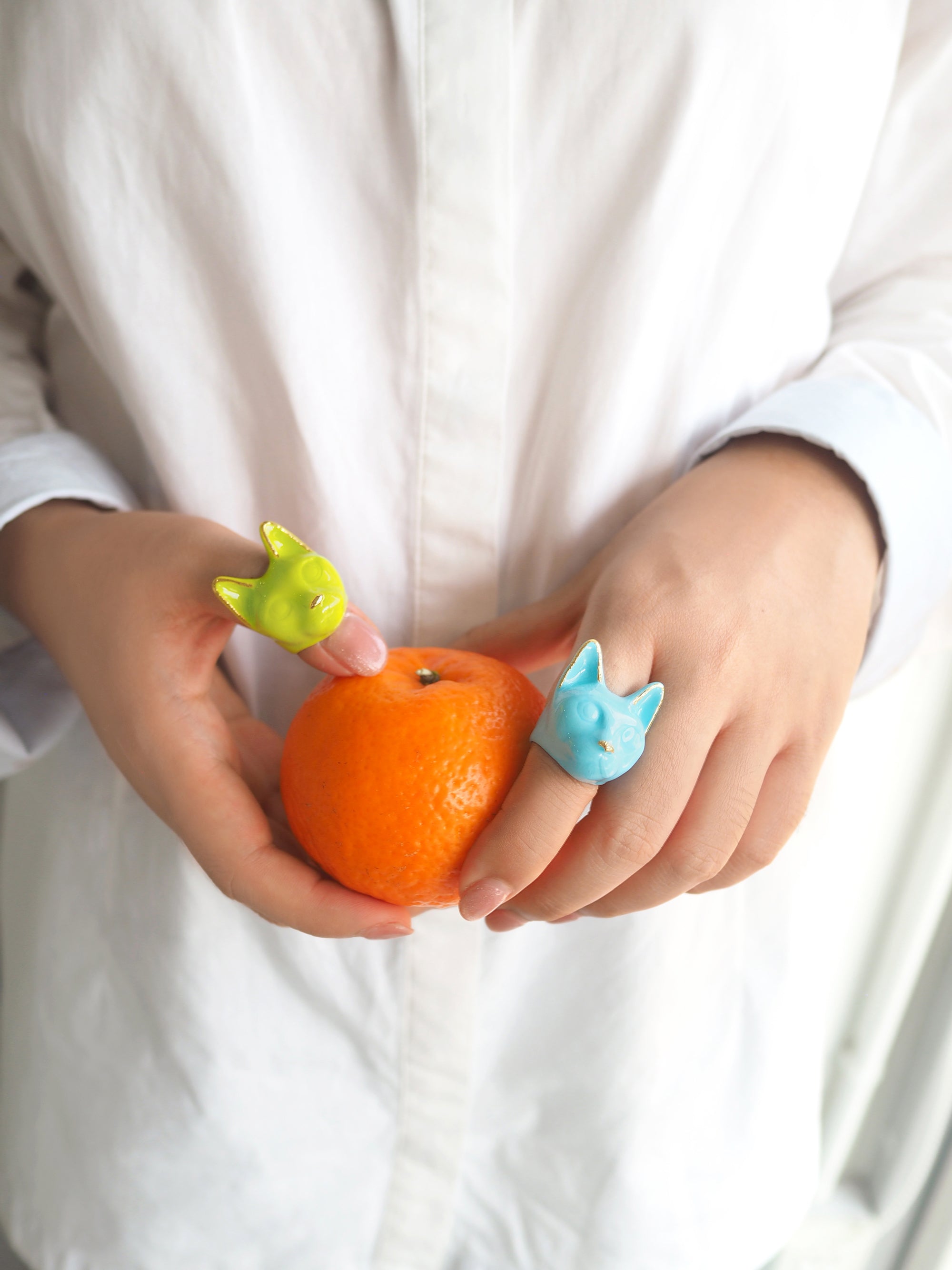 Two colorful cat rings, one green and one blue, on fingers holding an orange.