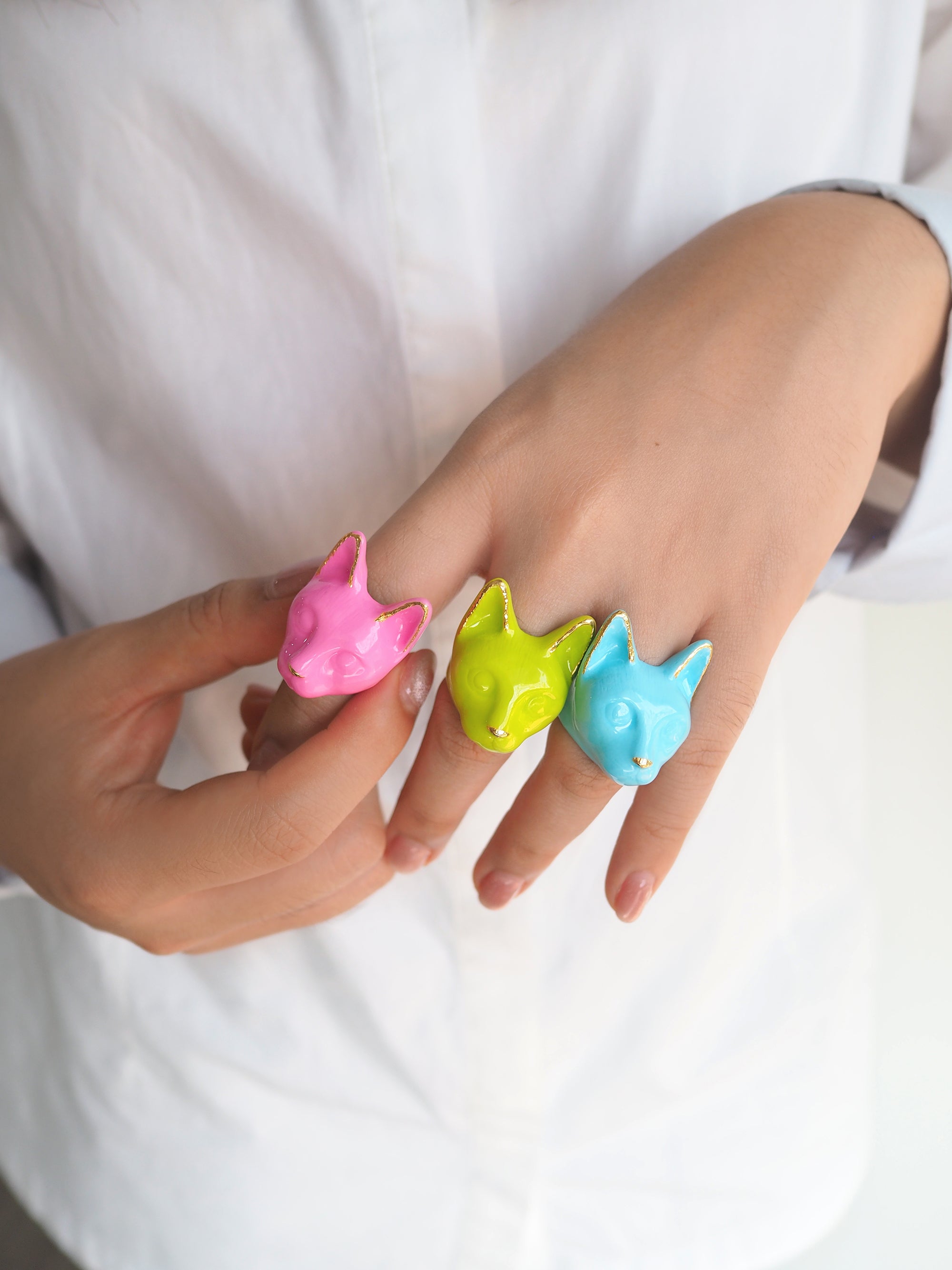 A hand wearing three colorful cat rings: pink, green, and blue, perfect for summer look.