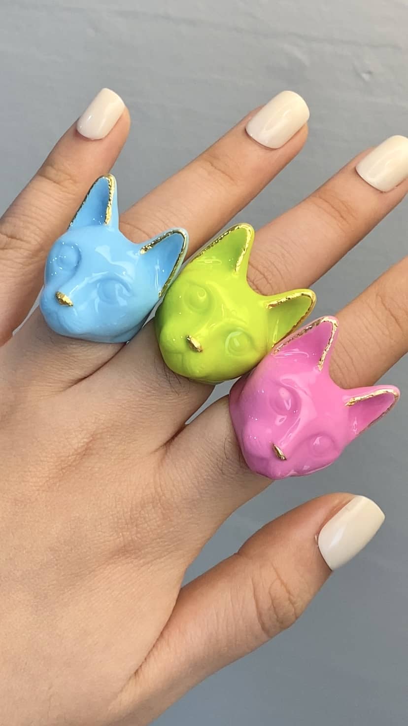 A hand wearing three colorful cat rings: pink, green, and blue, perfect for cat lovers.