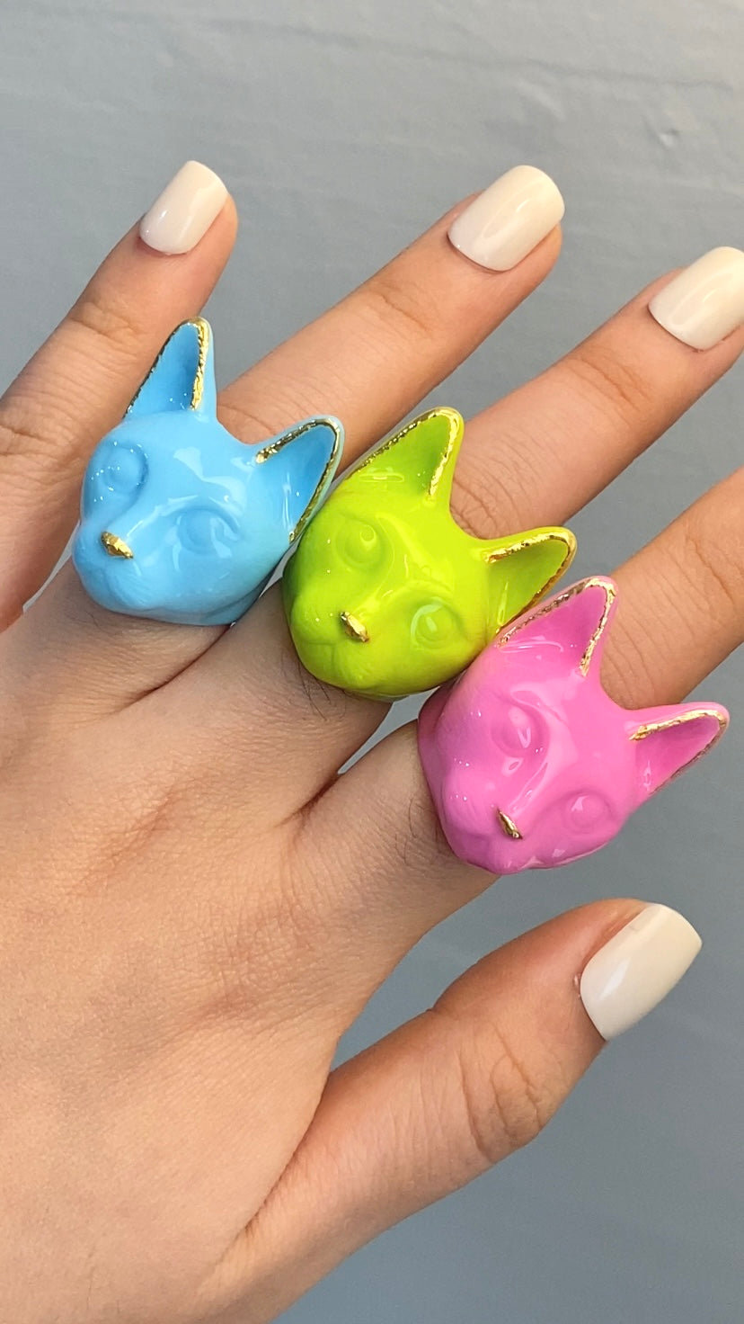 A hand wearing three colorful cat rings: pink, green, and blue, perfect for cat lovers.