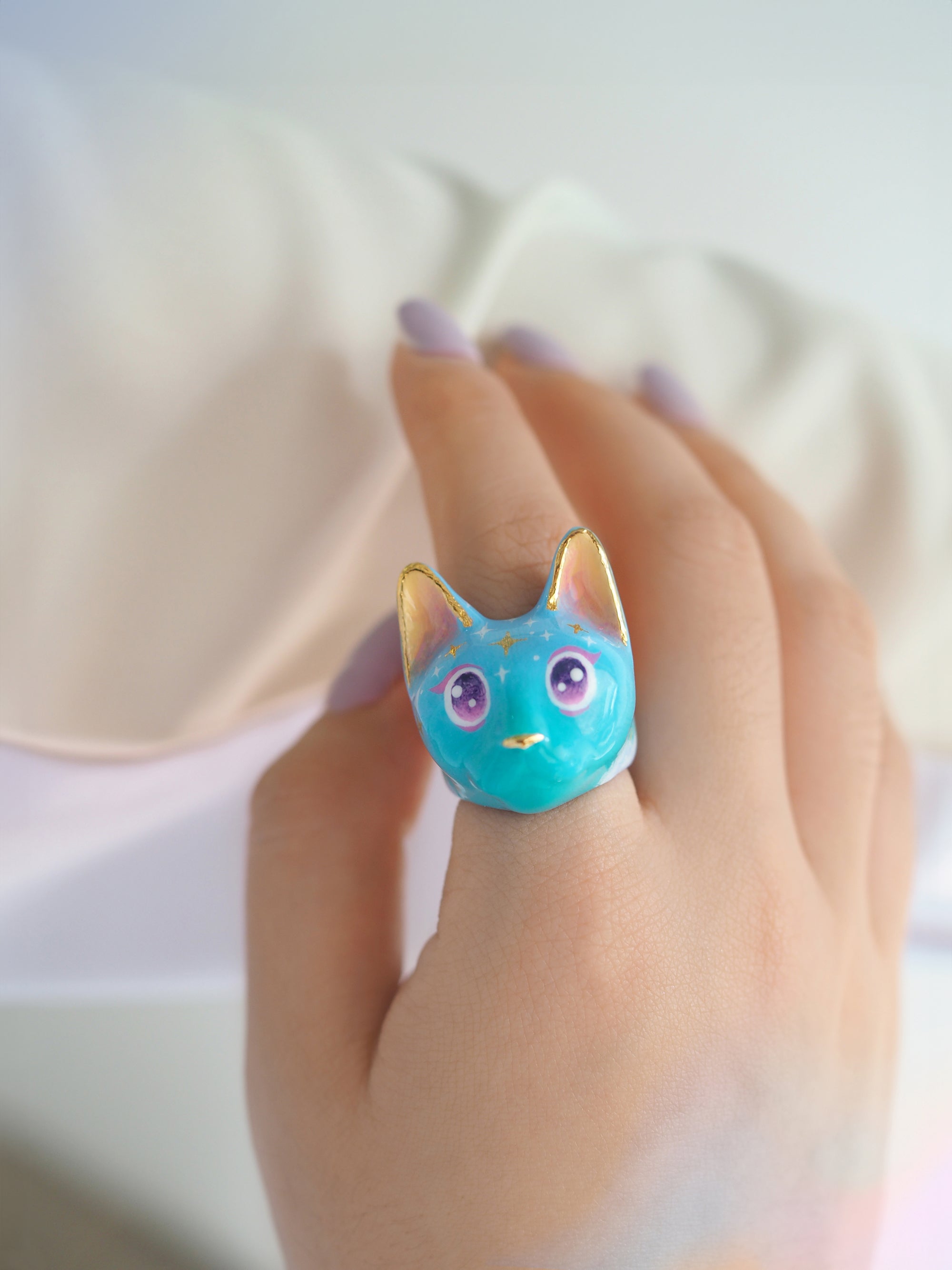 A close-up of a hand adored with a cat ring in a soft blue color featuring sparkling purple eyes and star-like patterns on its forehead.