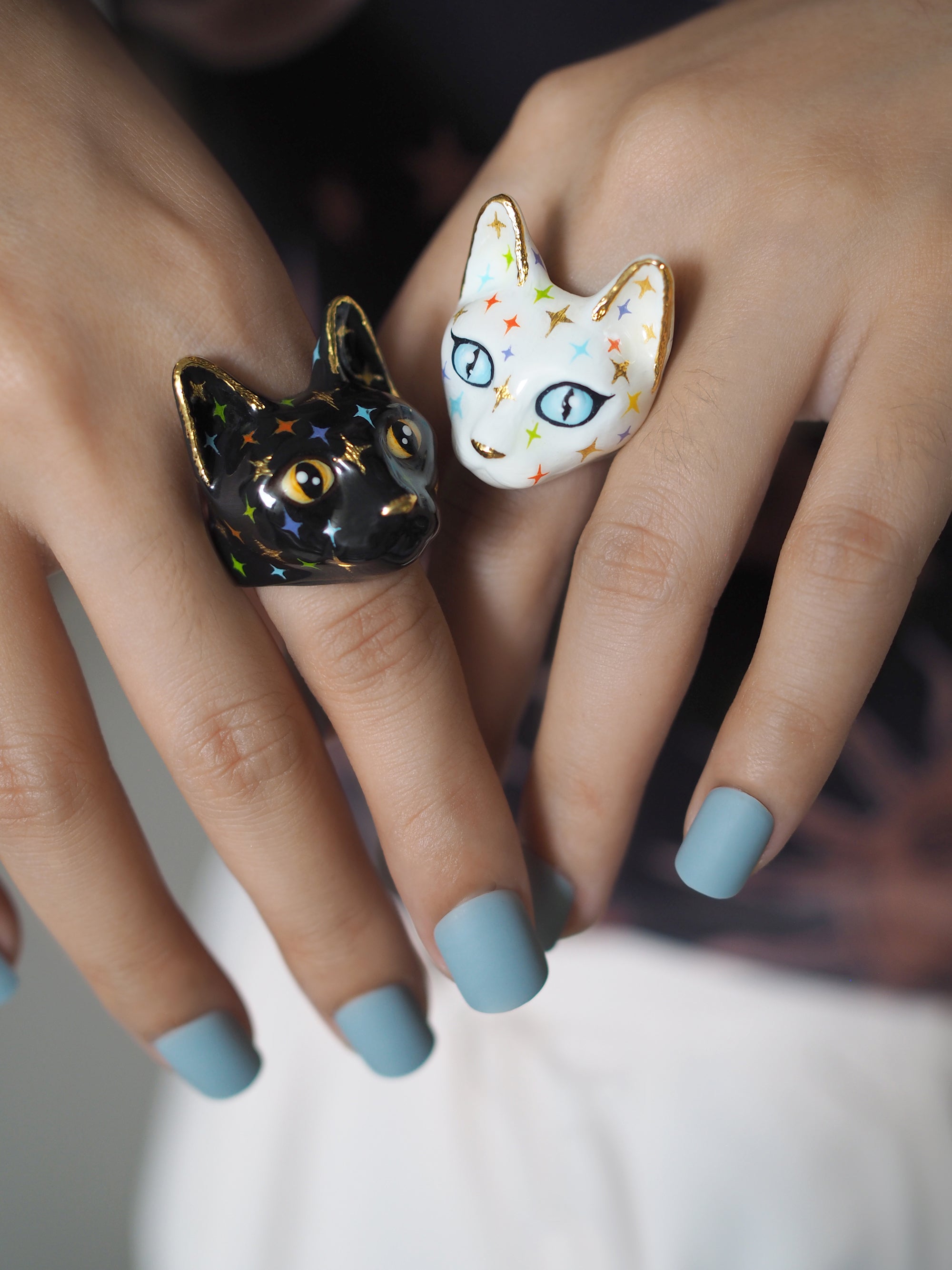 Hands wearing two cat rings: a black cat with yellow eyes and colorful twinkle details, and a white cat with blue eyes and twinkle details.