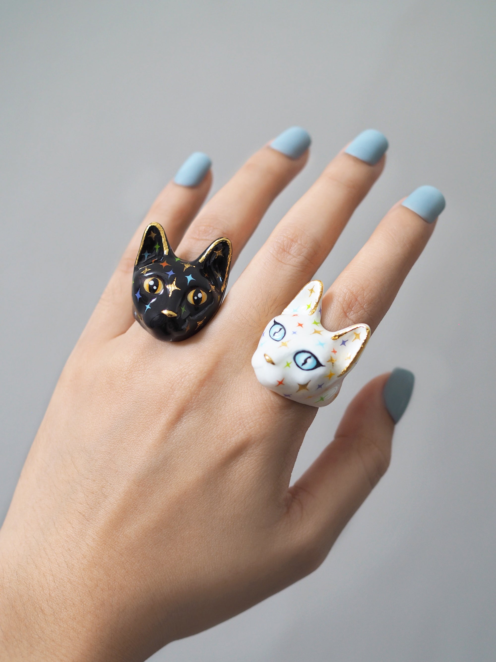 A hand with light blue nail polish wearing two cat face rings in white and black featuring a twinkle-shaped details on their faces.