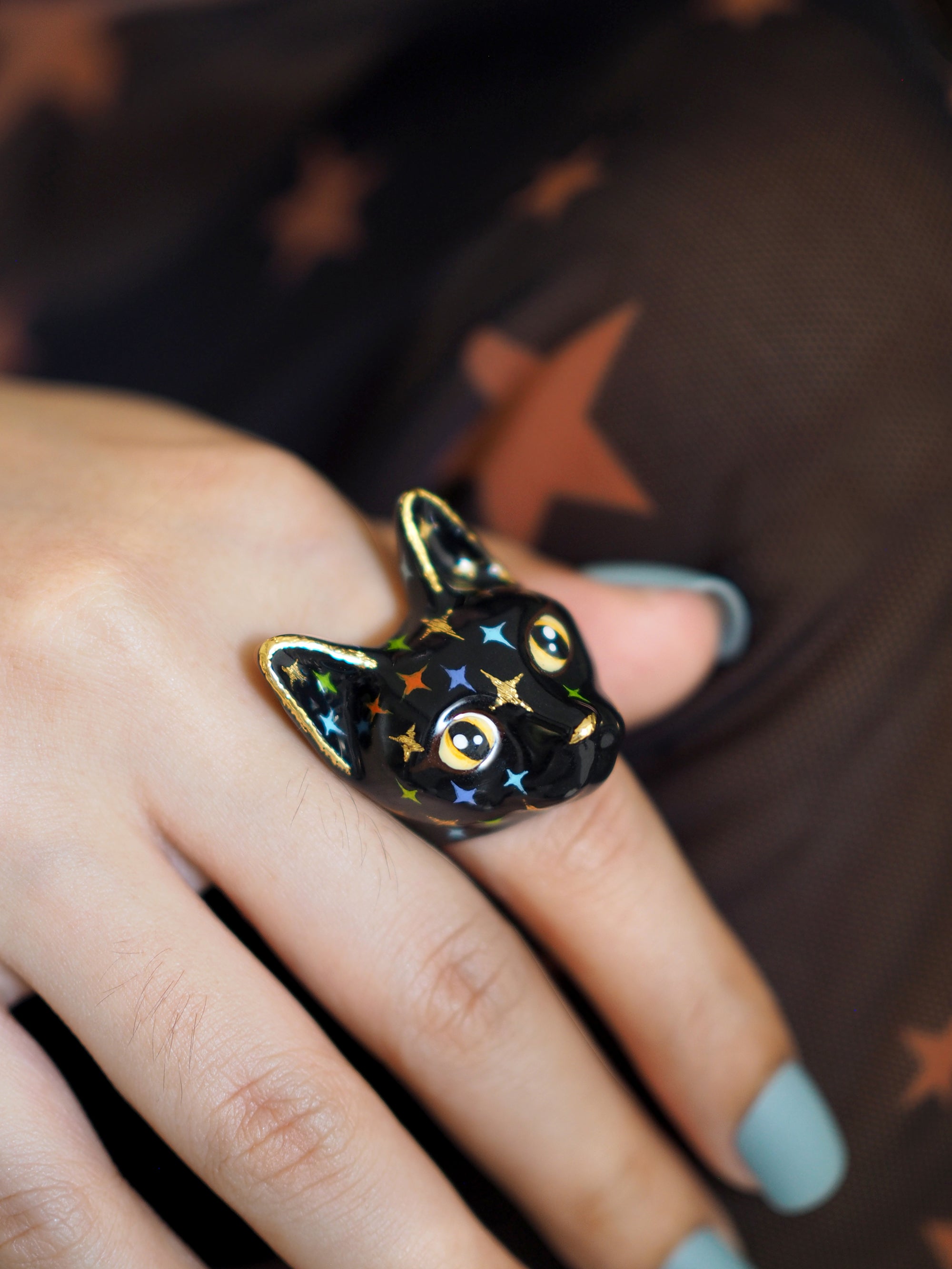 A hand wears a black cat ring with yellow eyes and a face full of colourful twinkle-shaped details.