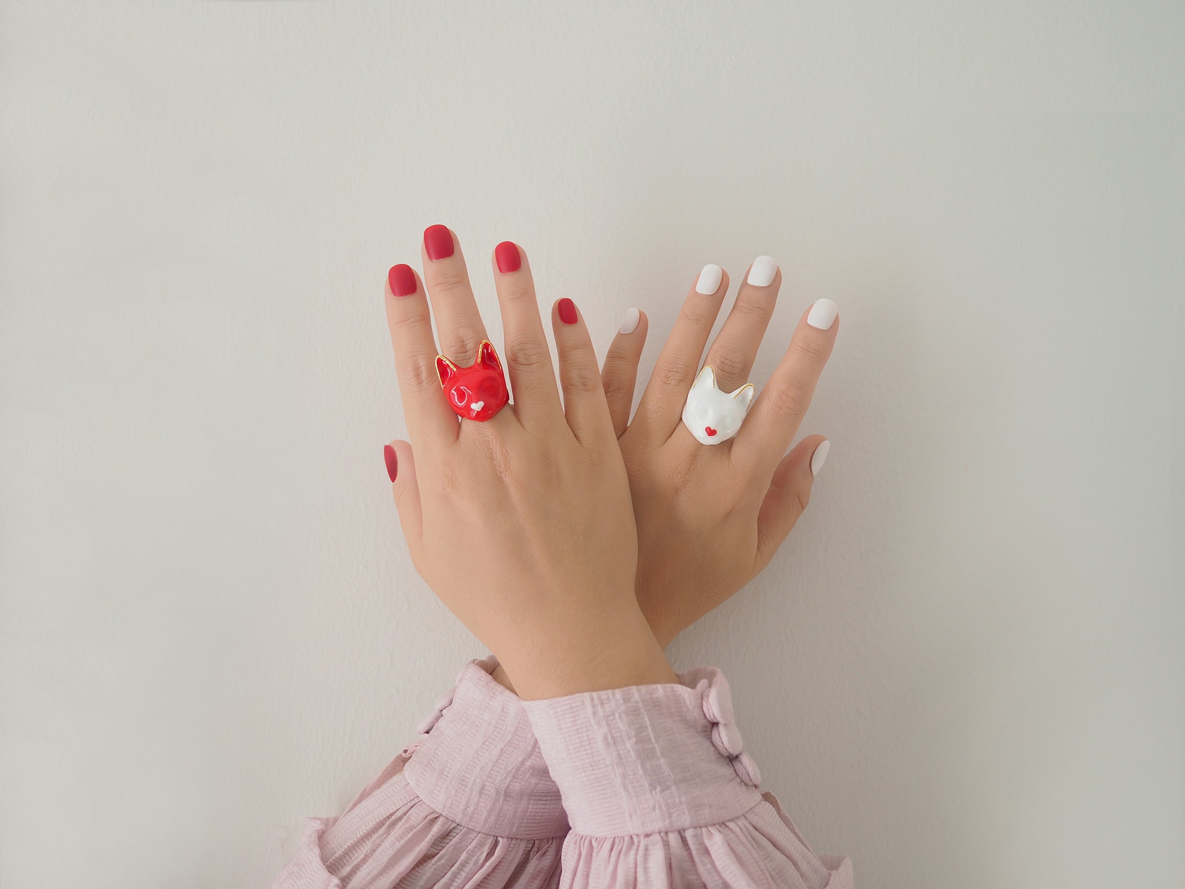 A close-up of two hands showcasing a white cat-shaped ring with a small red heart and a red cat-shaped ring with a small white heart against a plain background.