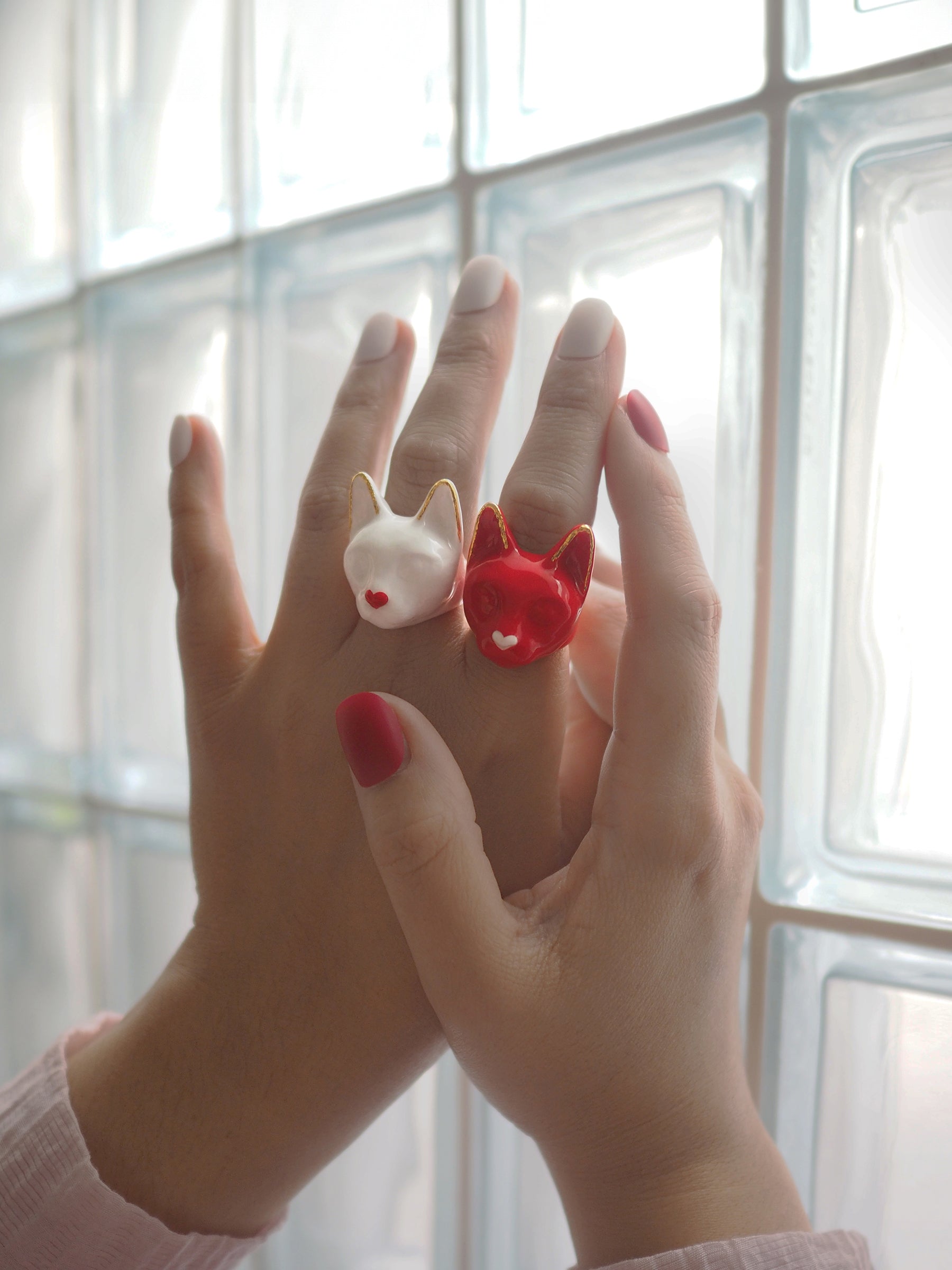 A close-up of a hand wearing a white cat-shaped ring with a small red heart and a red cat-shaped ring with a small white heart.