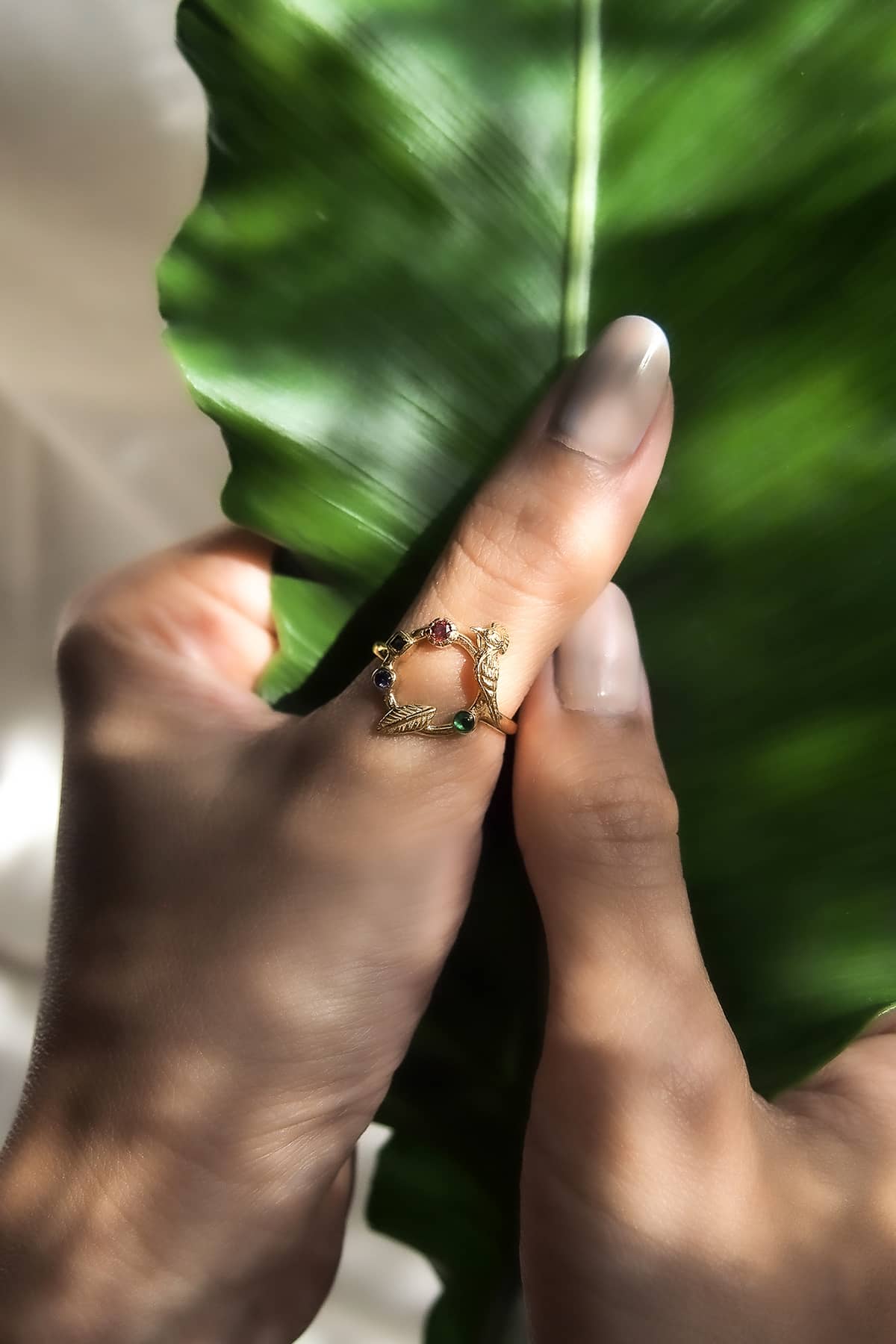 A close-up of a hand holding a green leaf, showcasing a gold ring featured bird design with colorful stones.