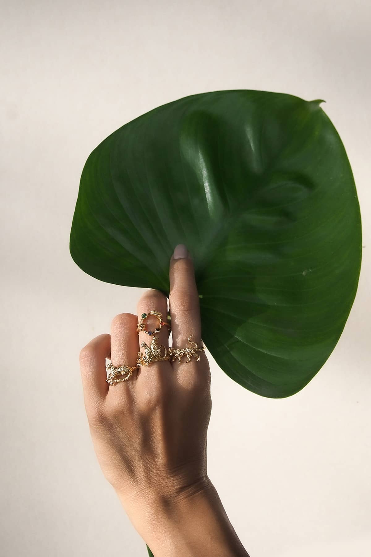 A hand displaying several nature-inspired gold rings, featuring leopard designs, holding a large green leaf against a neutral background.