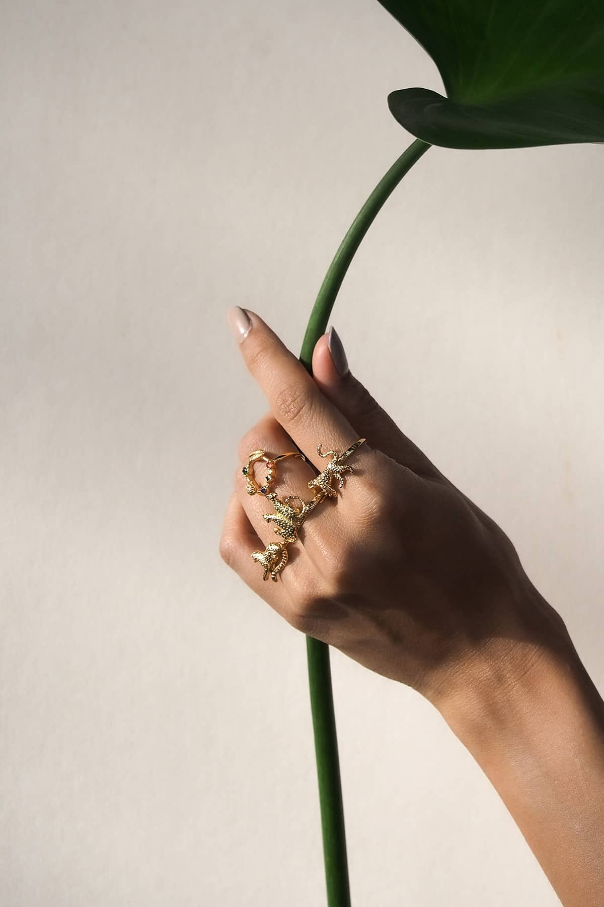 A hand displaying several nature-inspired gold rings, featuring leopard designs, holding a large green leaf against a neutral background.