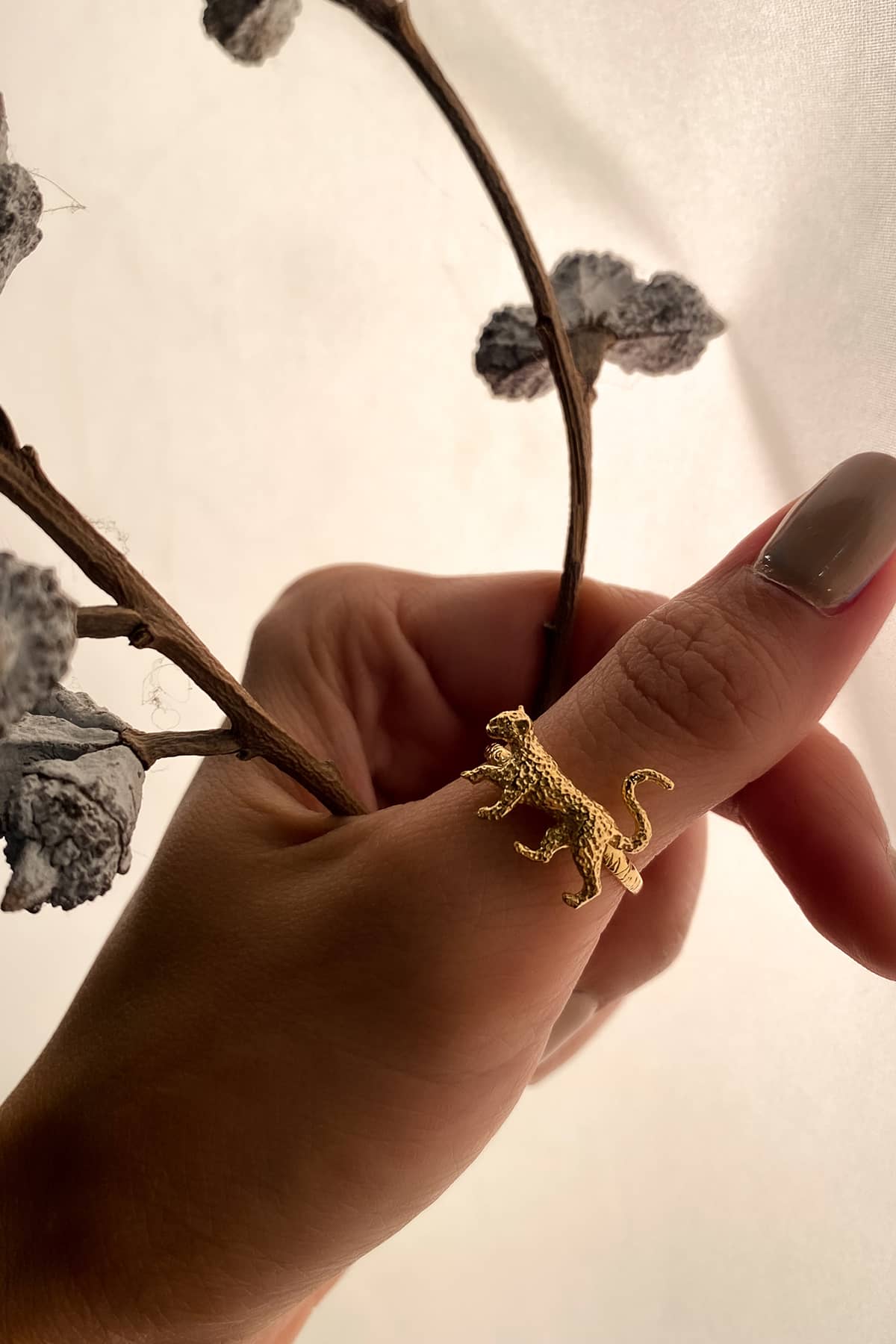 A hand with a neutral manicure displaying a gold leopard ring, surrounded by dried branches and leaves.