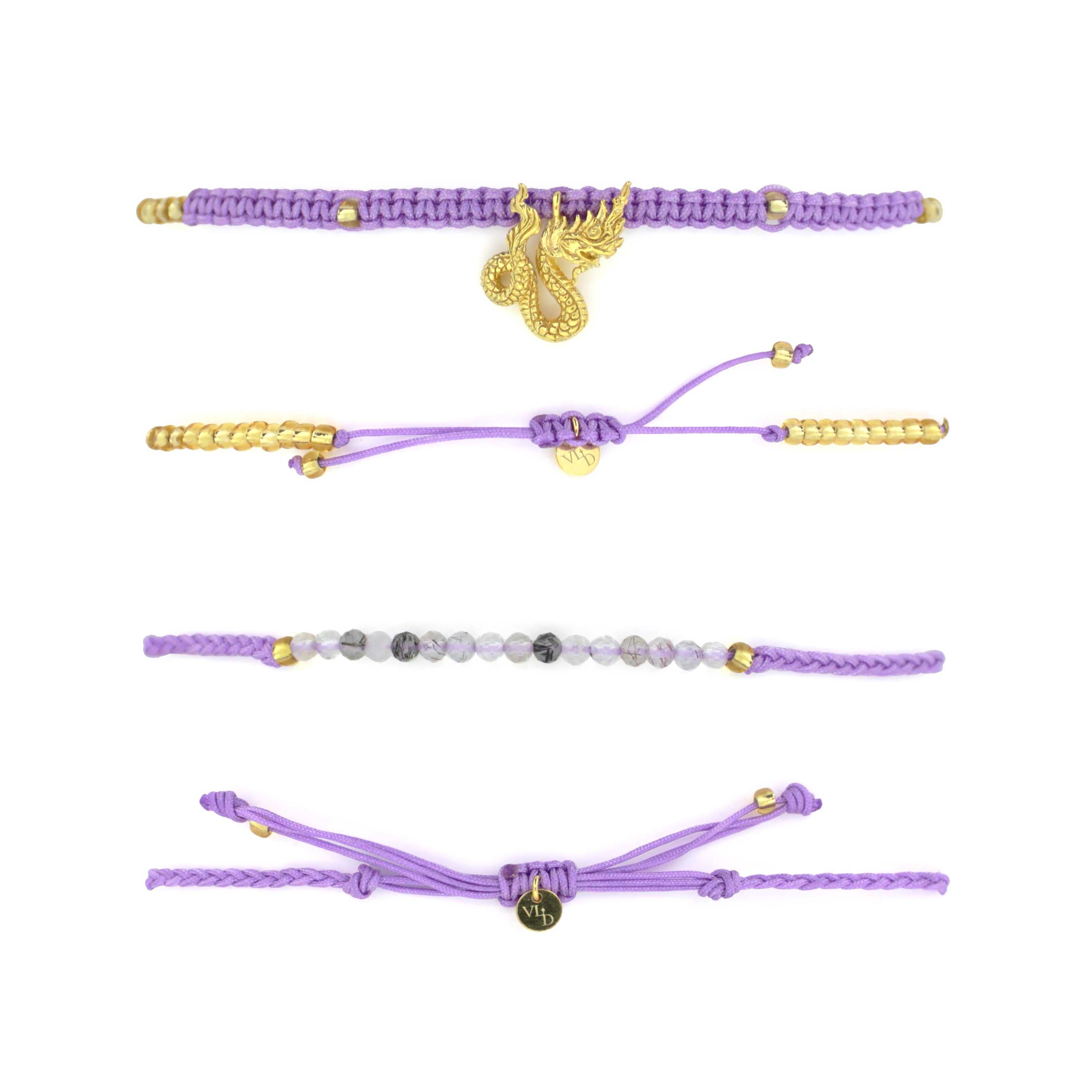 Set of two bracelets the top bracelet featuring a purple woven design material adorned with a gold Naga charm. The bottom bracelet features a series of grey and white beads with a simple woven purple band.