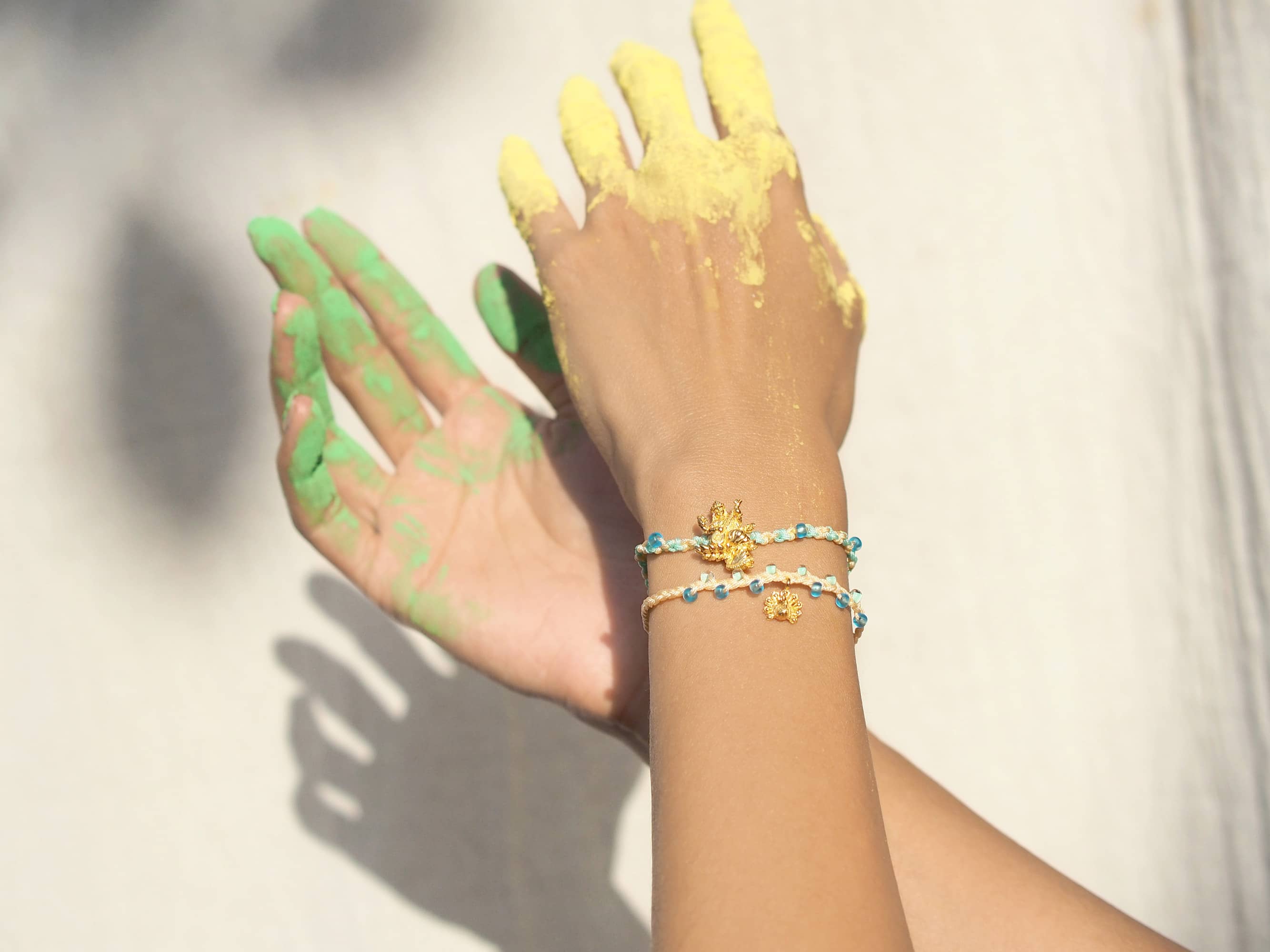 A hand adored with two pastel bracelets featuring a gold Tridevi Sarasvati charm and a gold peacock charm.