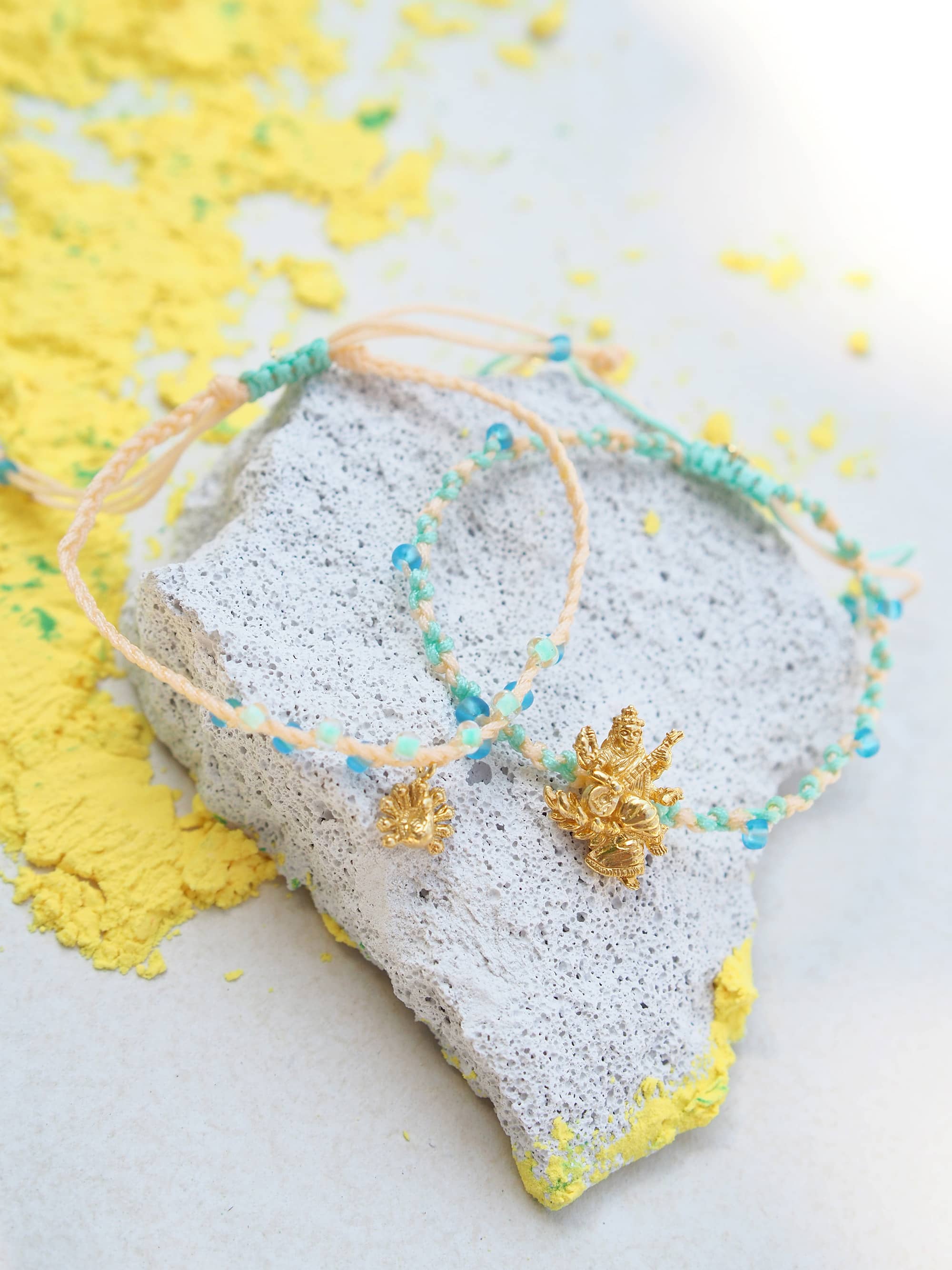 Close-up of two bracelet featuring a gold Tridevi Sarasvati charm and a gold peacock charm placed a textured surface next to yellow powder.