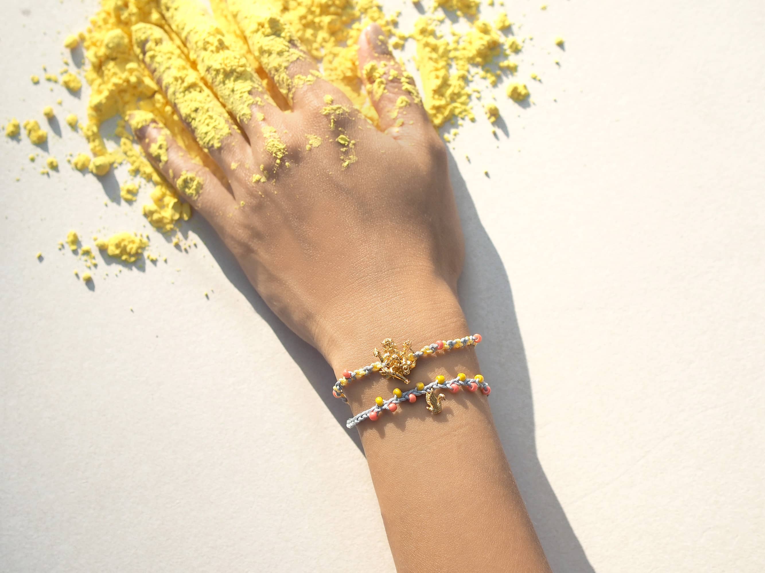 A hand with yellow powder covering its fingers, while wearing charm bracelet featuring Trimurti Brahma and swan charms.