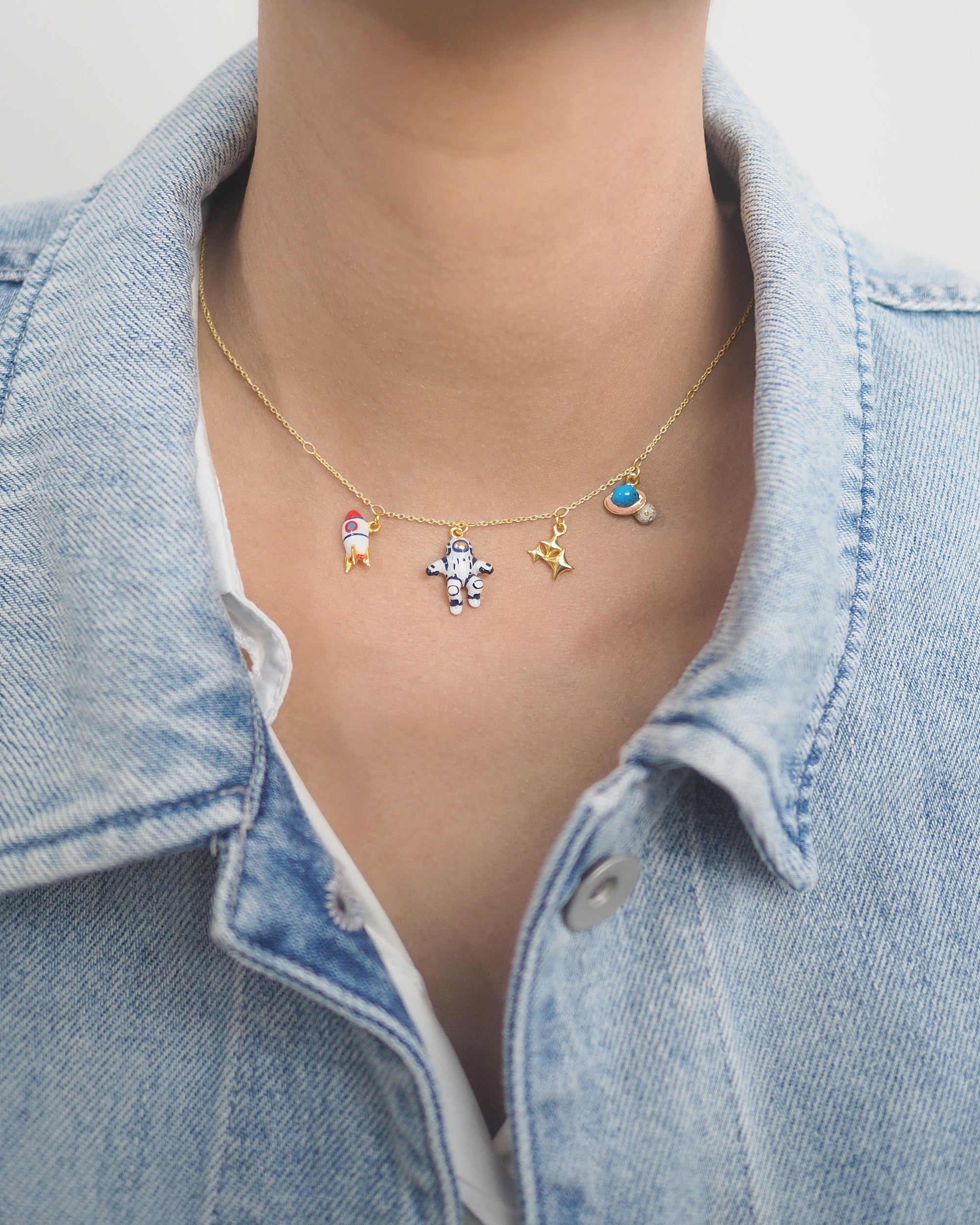 Close-up of a person wearing a denim jacket and a gold necklace with colorful charms: a rocket, astronaut, stars, and Saturn charms