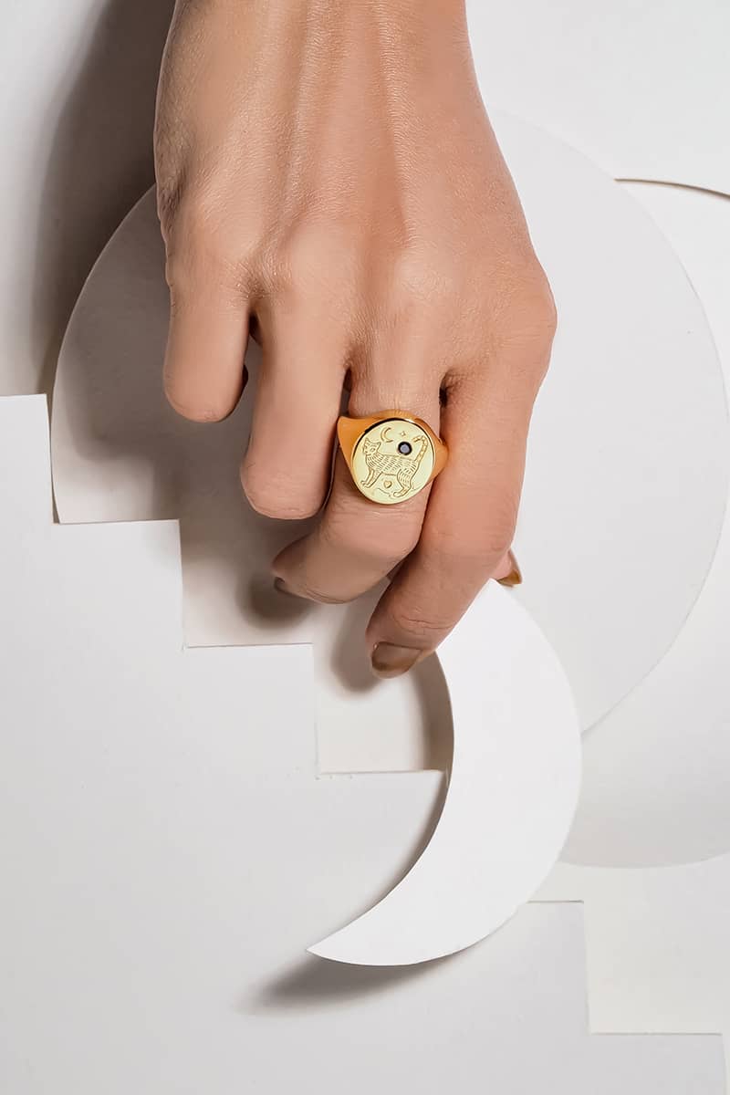 A beautiful gold signet ring featuring an engraved cat design with a synthetic black gemstone and moon motifs is worn on a hand while holding a moonlike-shaped paper.