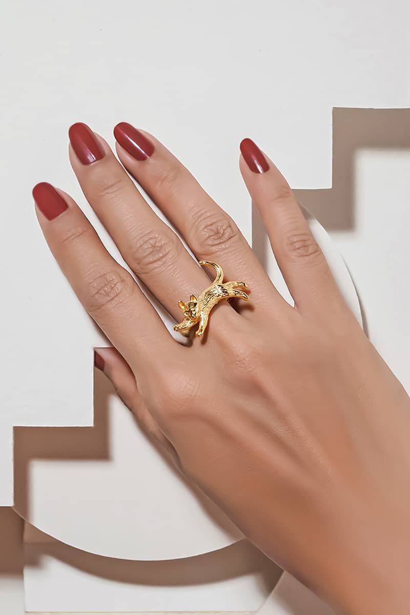 Close up of a hand with red nail polish wearing a gold ring featuring a adorable jumping cat design.