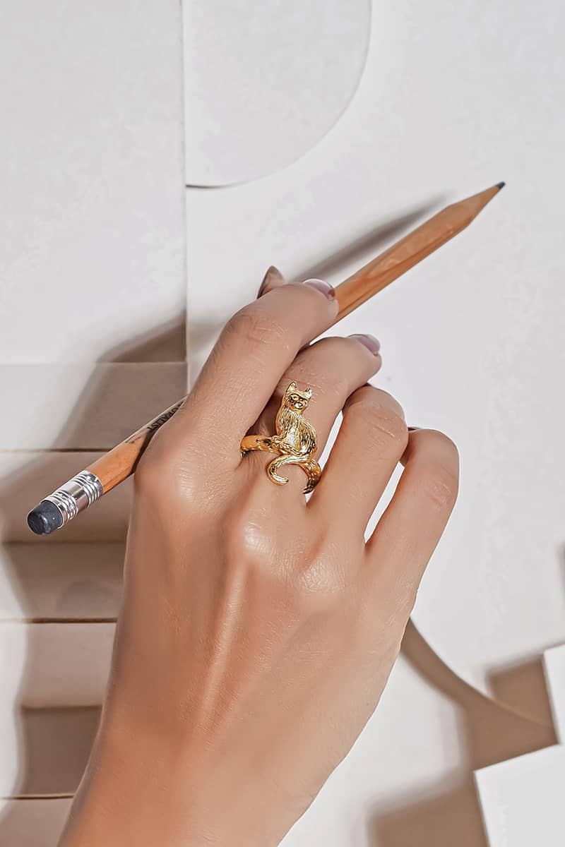 A hand adorned with a gold ring featuring setting cat design while holding a pencil.